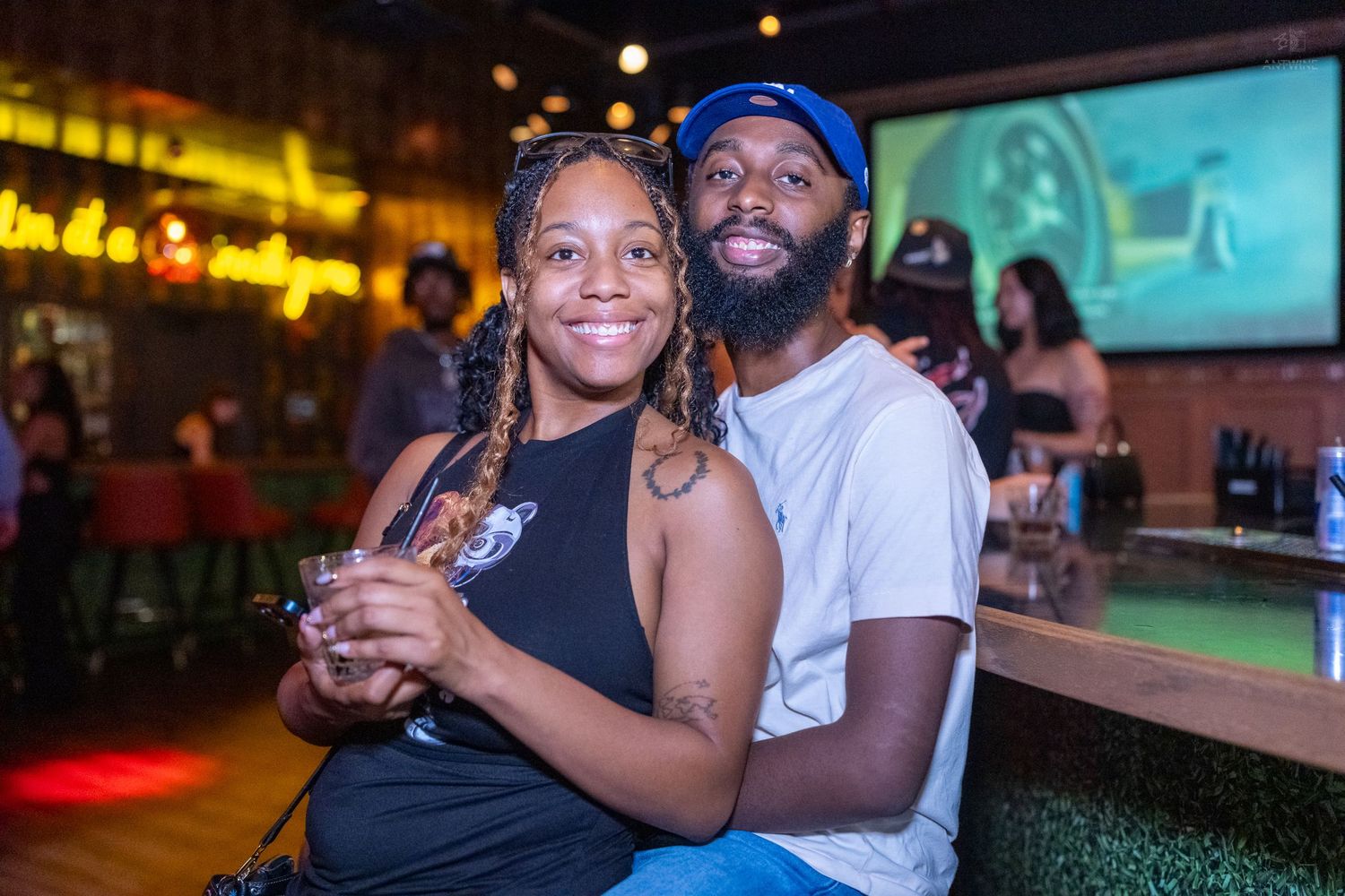A smiling couple poses together at a bar, with neon signs and a screen visible behind them.