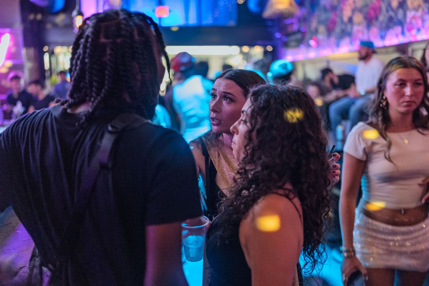 Young women conversing on a crowded purple-lit nightclub dance floor.
