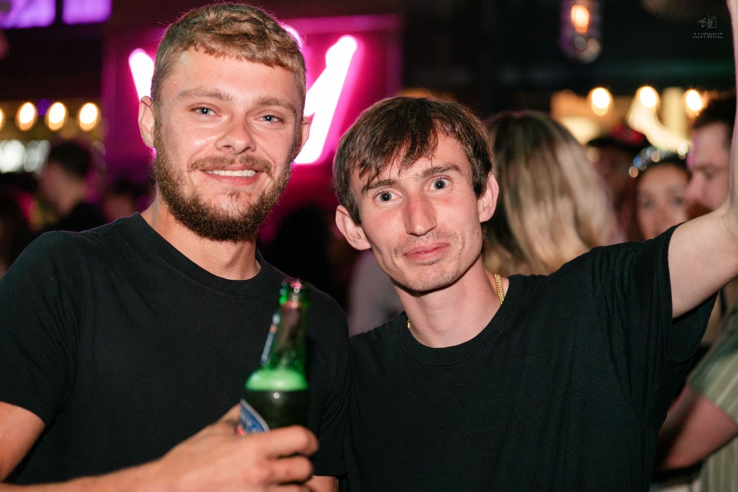 Two men in black t-shirts smiling at a bar, one holding a green beer bottle under neon lights.