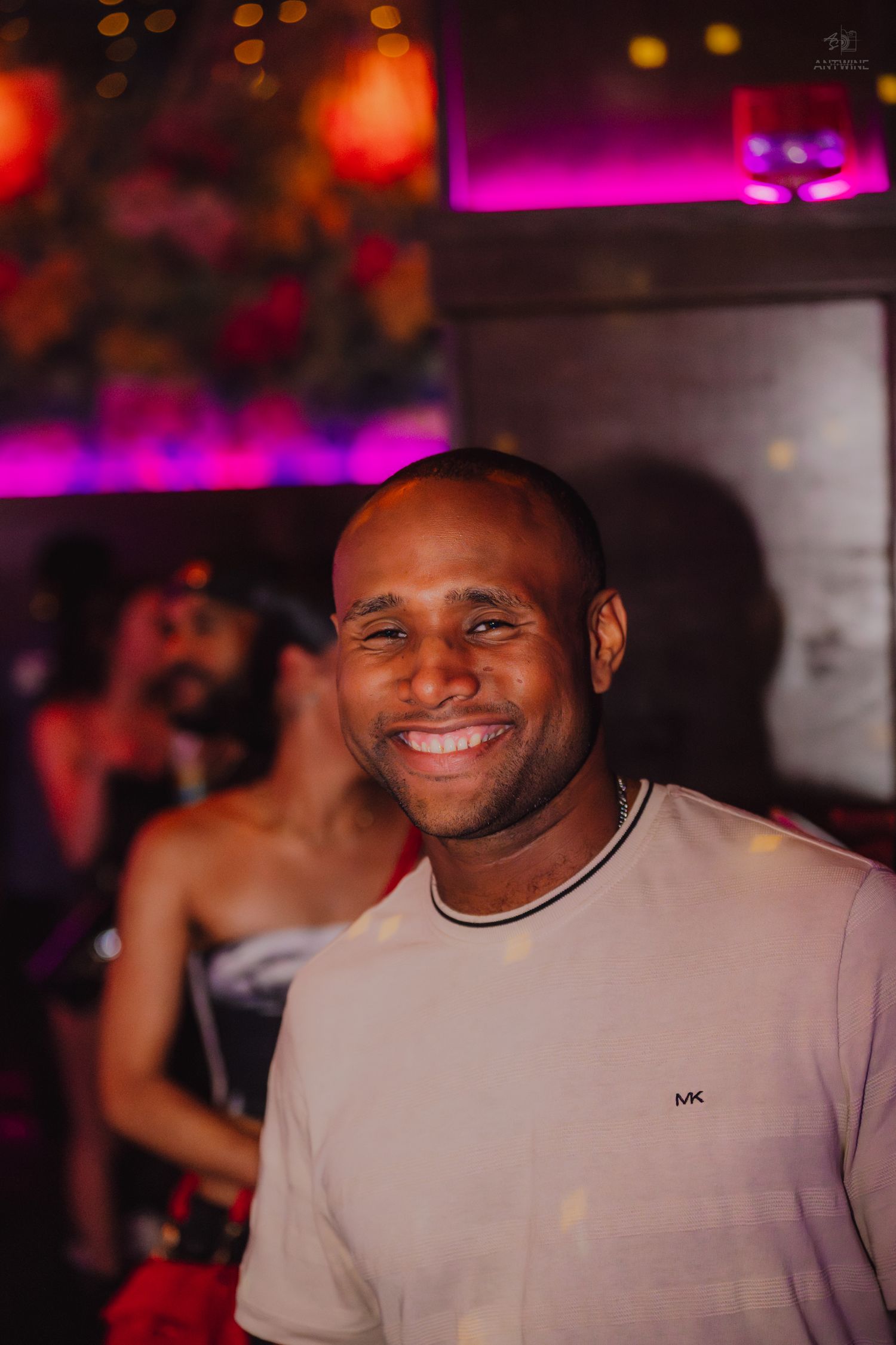 A smiling bald Black man wearing a Michael Kors cream t-shirt poses in a vibrant nightclub setting.