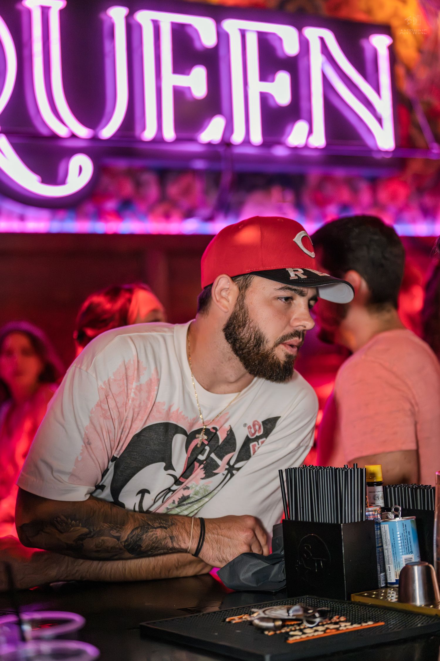 Bearded man in red Cincinnati cap and tattoos leans on bar under neon Queen sign at nightclub.