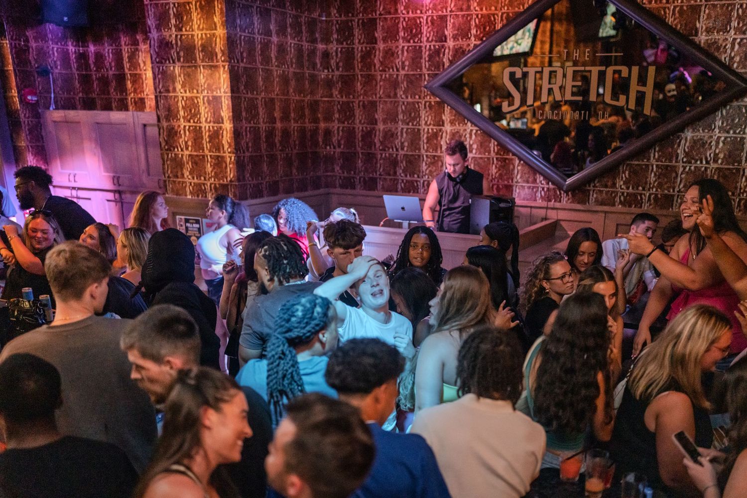 Energetic crowd dancing beneath a diamond-shaped Stretch nightclub sign with vibrant blue and purple lighting.