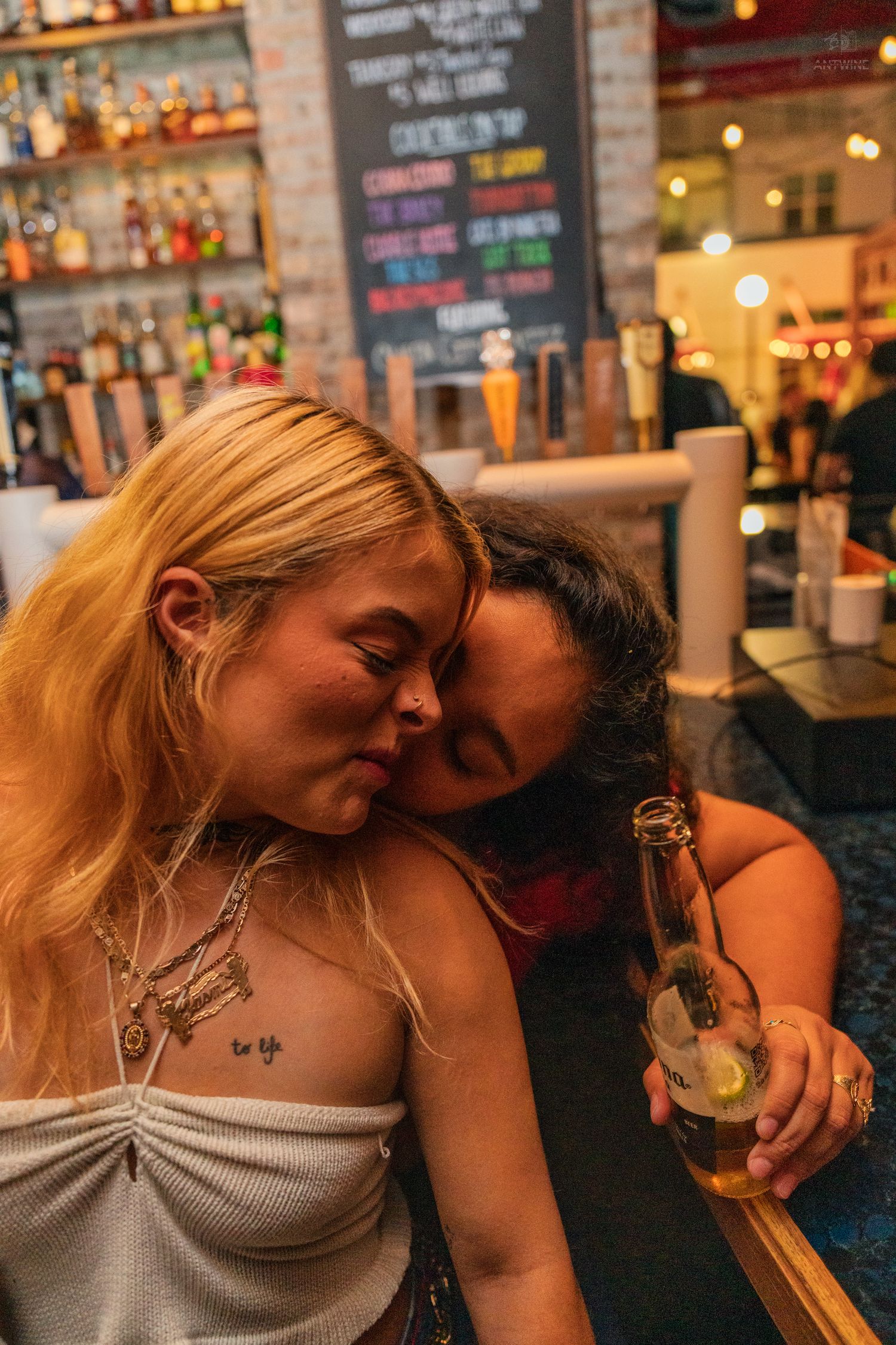 Two women cuddle close together at a bar, one holding a beer bottle with a lemon slice.