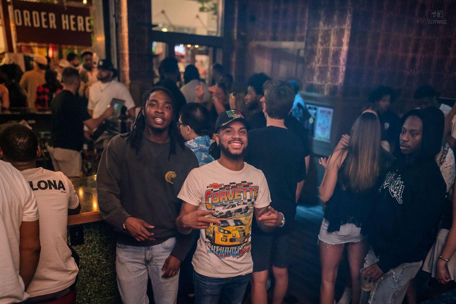Two smiling men posing together in a busy nightclub with Order Here sign in background.