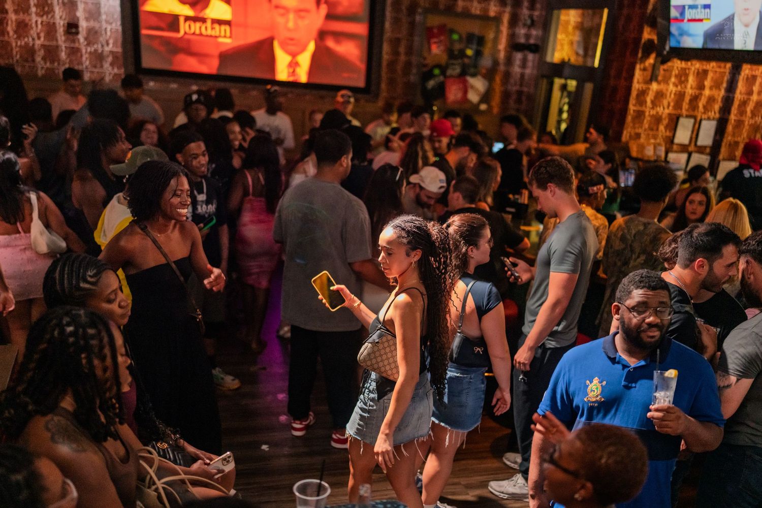 Busy bar crowd with people holding drinks, phone, TV screen visible in background.