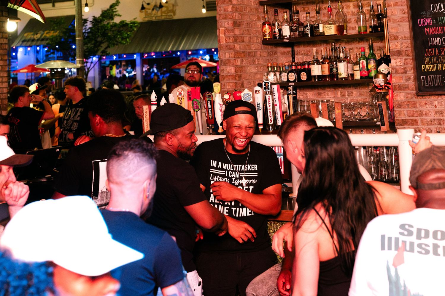Lively bar scene with diverse crowd socializing near a well-stocked liquor shelf at a busy nightlife venue.