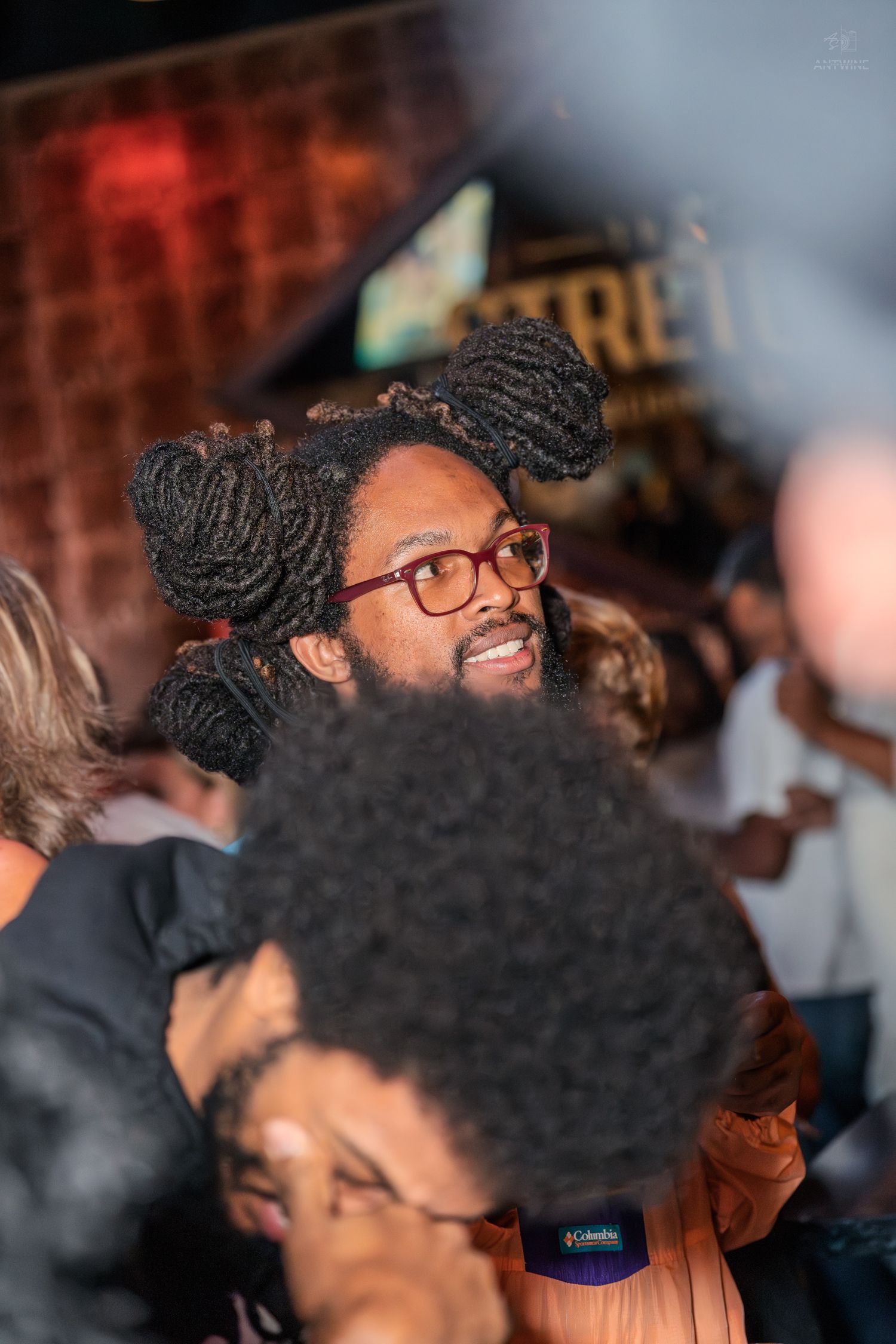 Man with styled dreadlocks and red glasses looking up in a crowded nightclub setting.