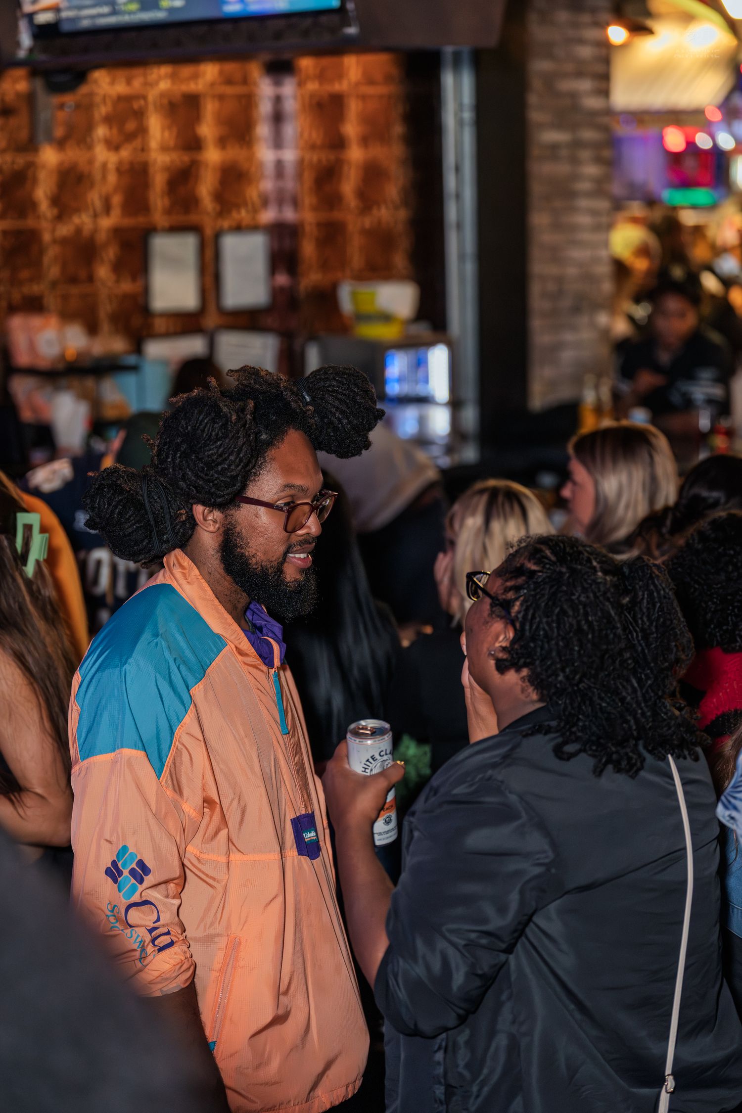 Man in orange Columbia jacket chatting with woman at busy bar.