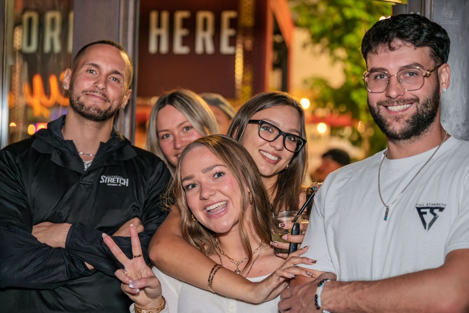 Five friends smile and pose together outside a venue with a HERE neon sign.