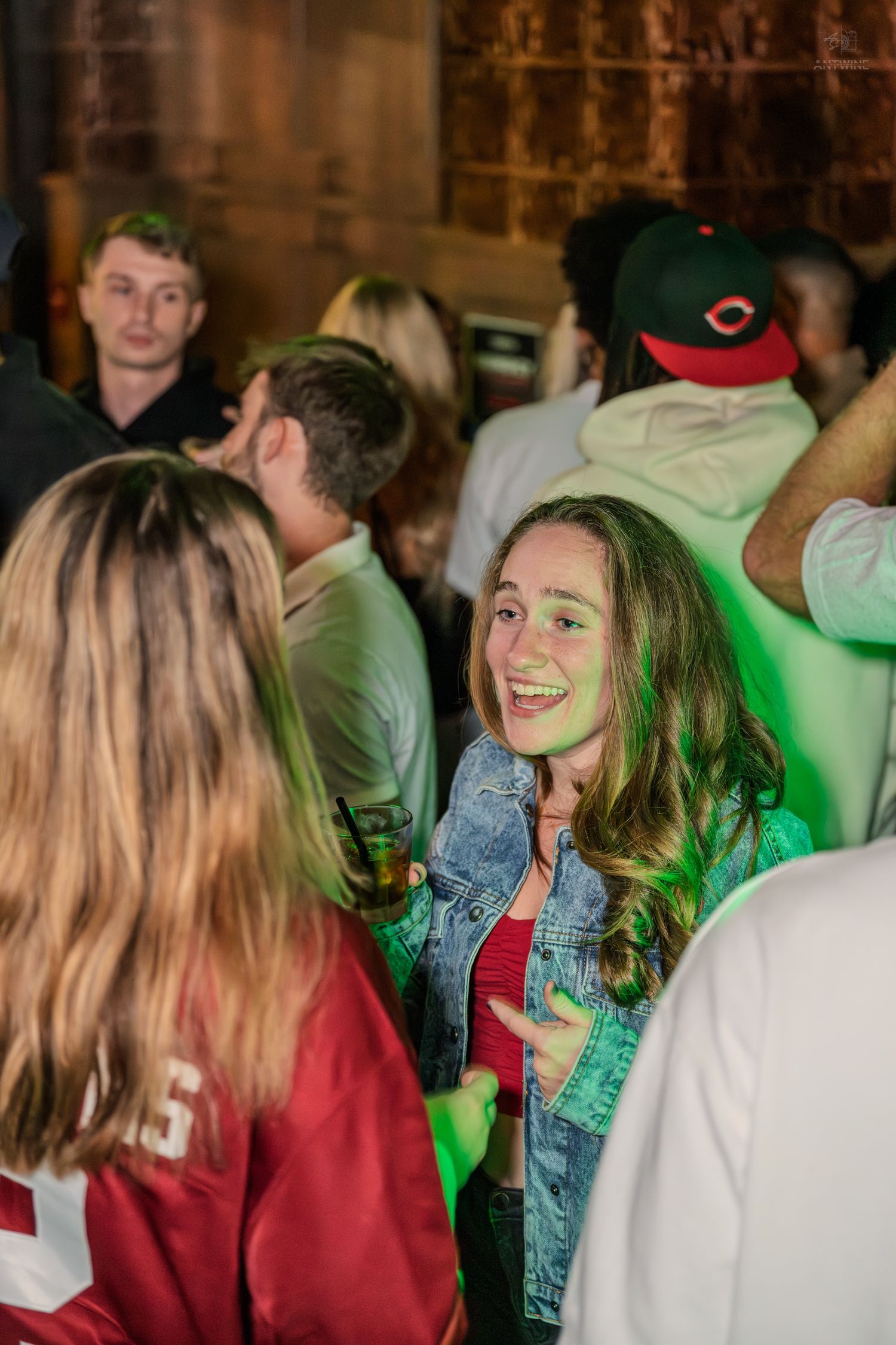 Happy young woman in denim jacket smiling at a lively nightclub party with green lights.