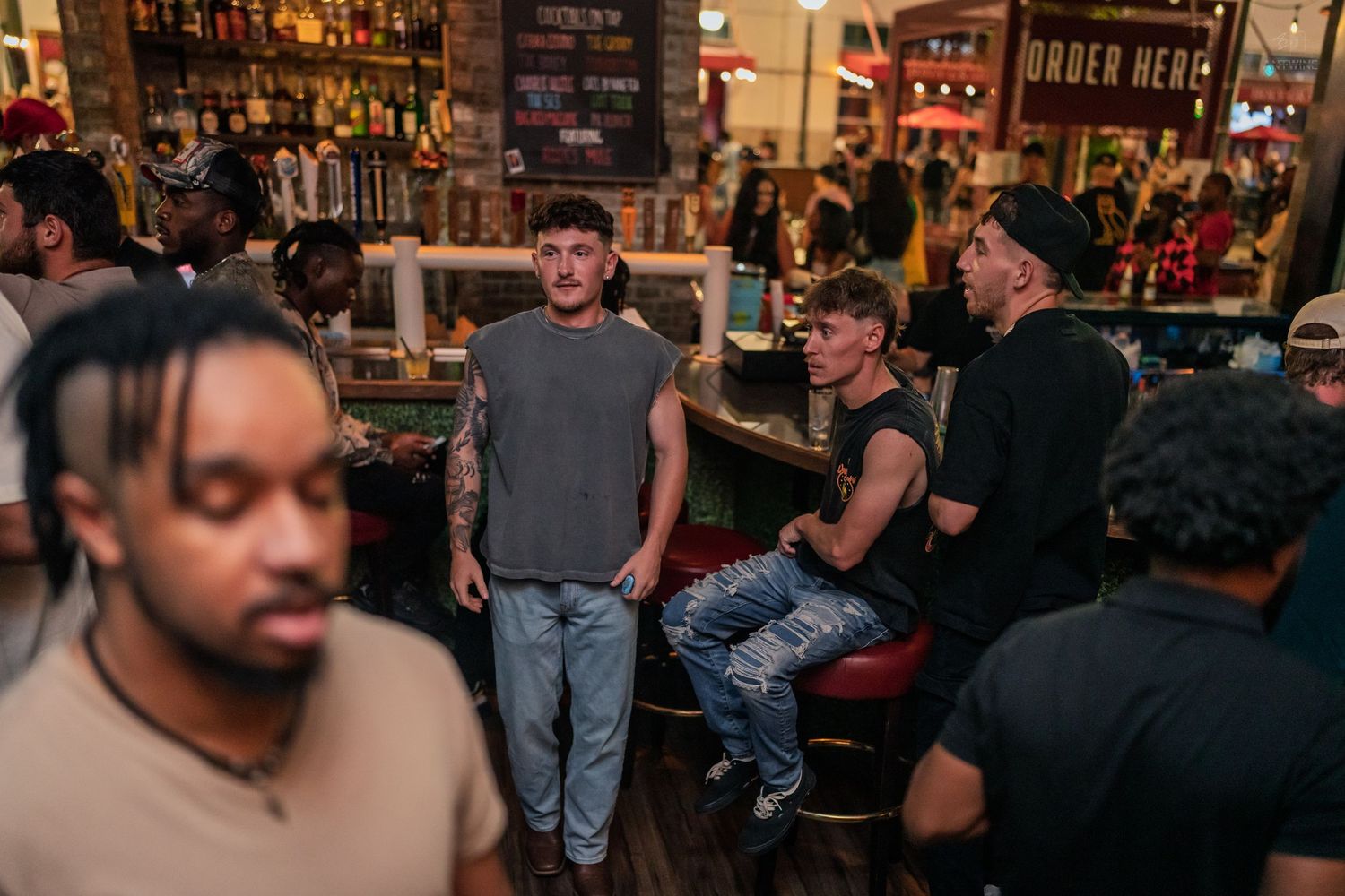 Group of young men socialize at a busy bar counter with shelves of liquor bottles in the background.