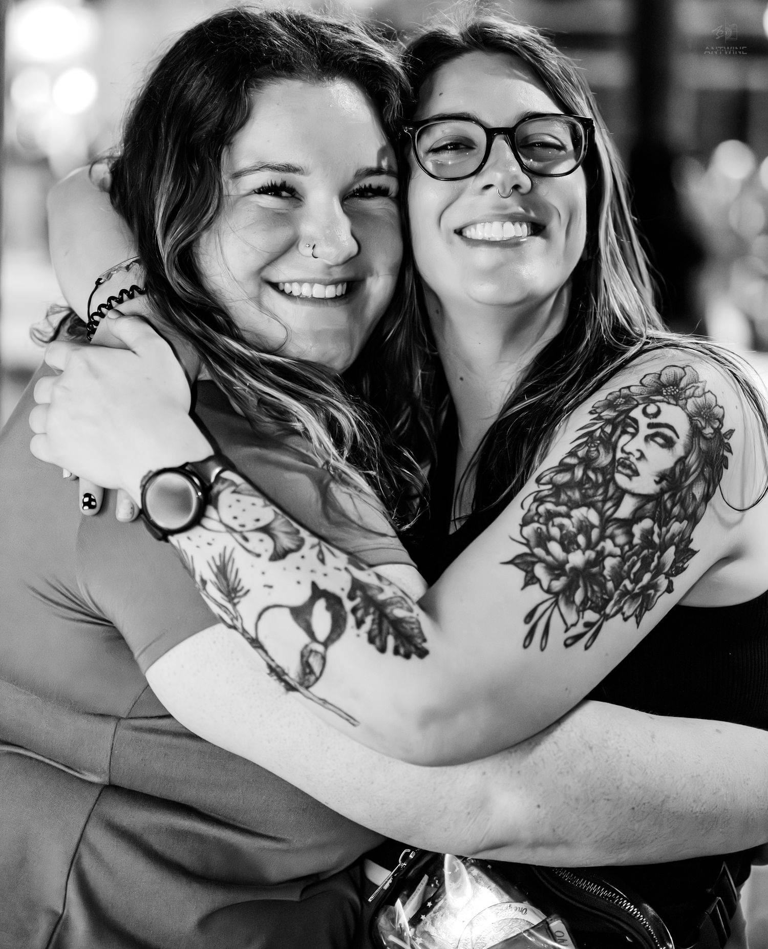 Two smiling women embracing, showing detailed floral and portrait arm tattoos in black and white.