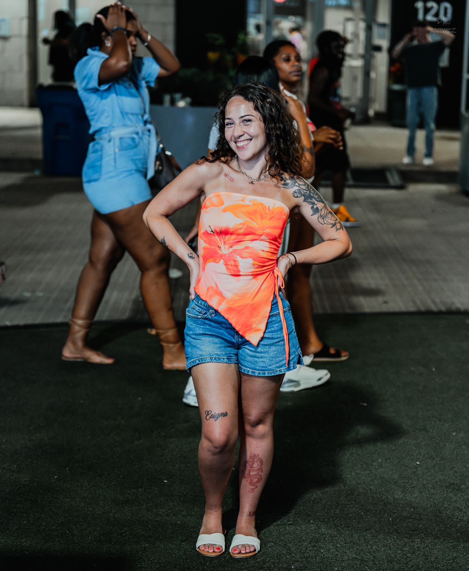 A smiling tattooed woman in an orange top and denim shorts poses confidently at an outdoor nighttime event.