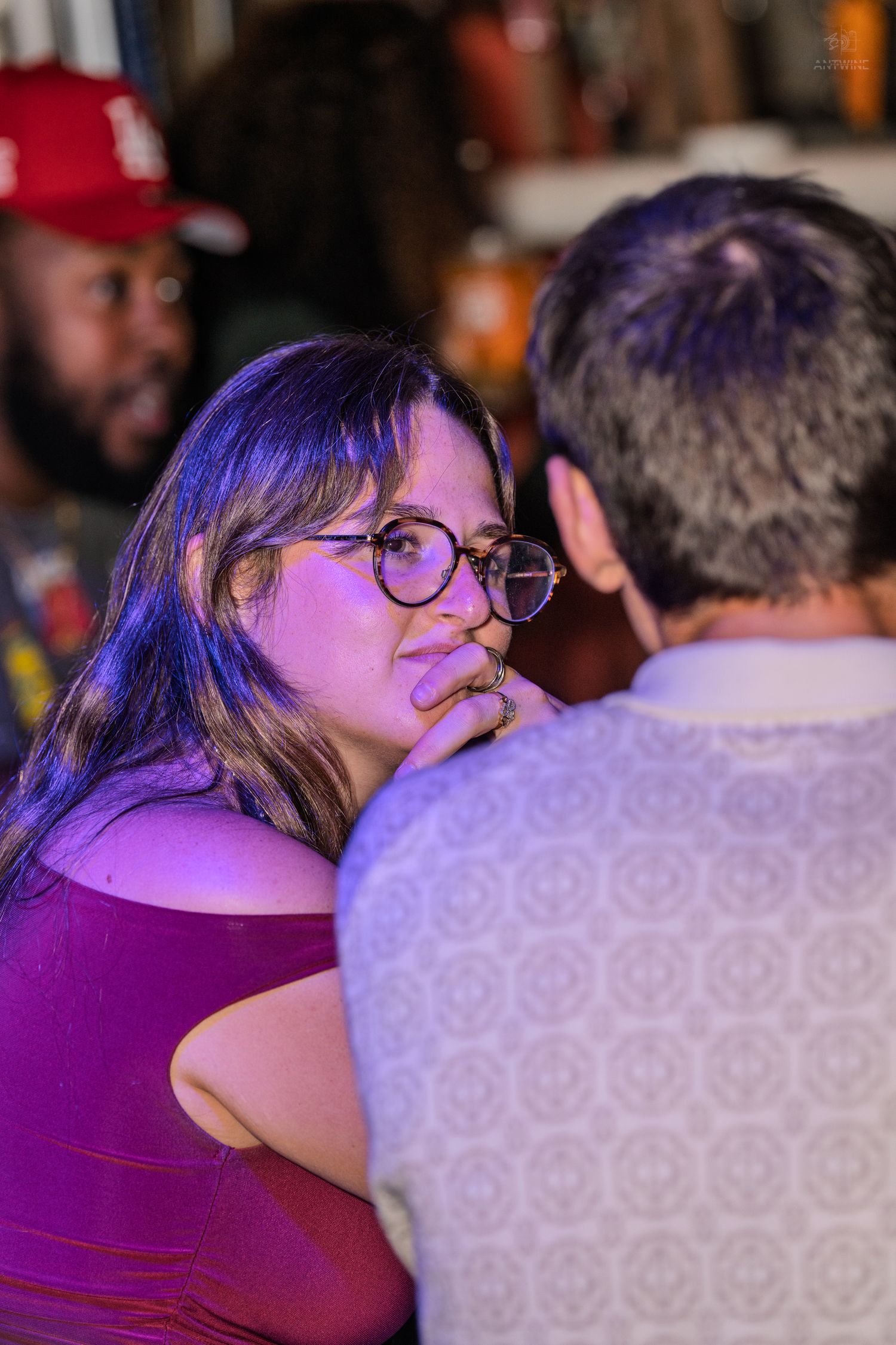Woman with glasses in a purple dress listens intently to a man at a bar.