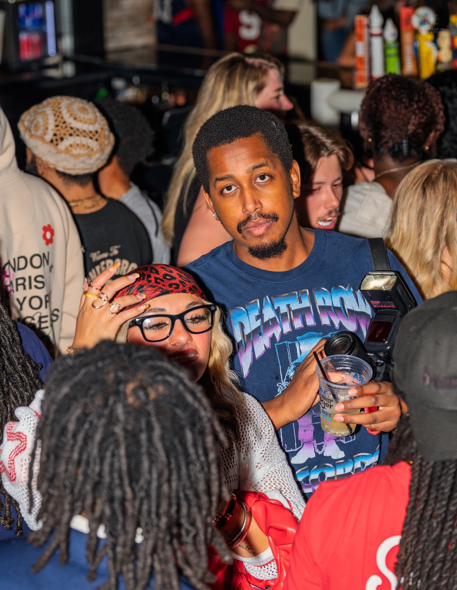 Man in Death Row Records shirt holding drink mingles in a busy bar crowd.