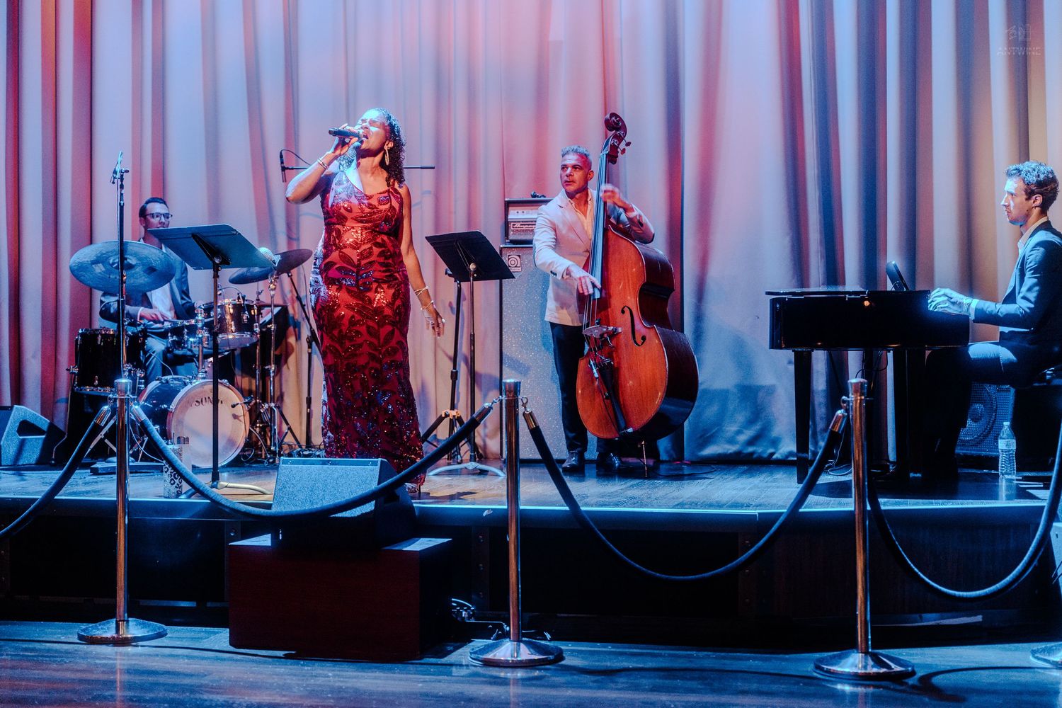 Elegant female jazz vocalist in red gown performing with a trio on a cruise ship stage.