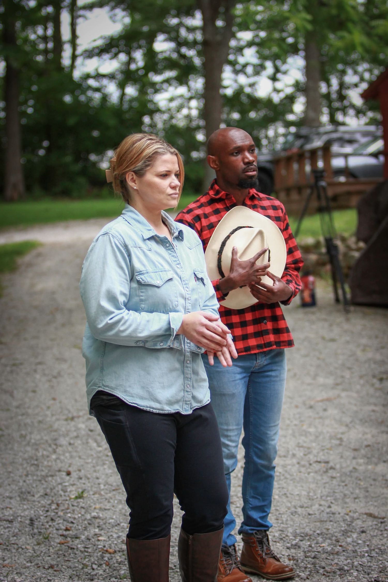 Woman in denim shirt and man in red plaid holding cowboy hat stand on gravel path outdoors.