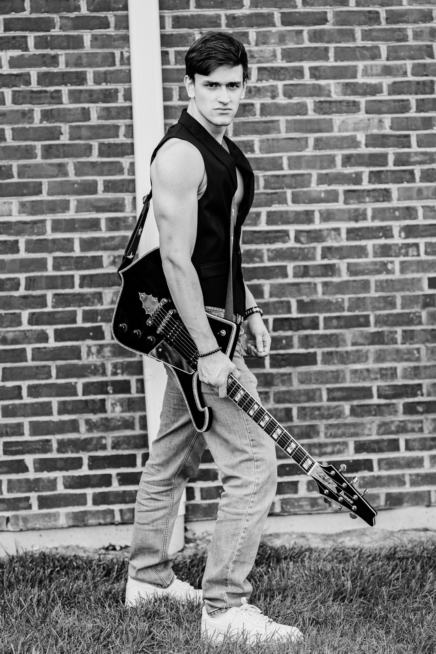 Black and white photo of young male musician posing with electric guitar by brick wall.