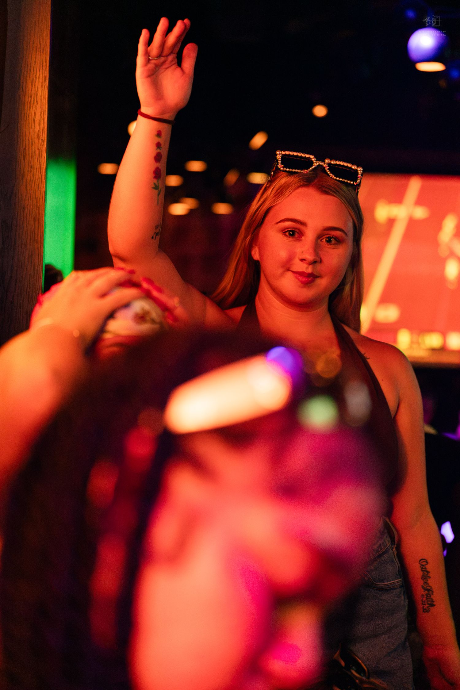 Smiling blonde woman with sunglasses raising her hand at a vibrant nightclub event.