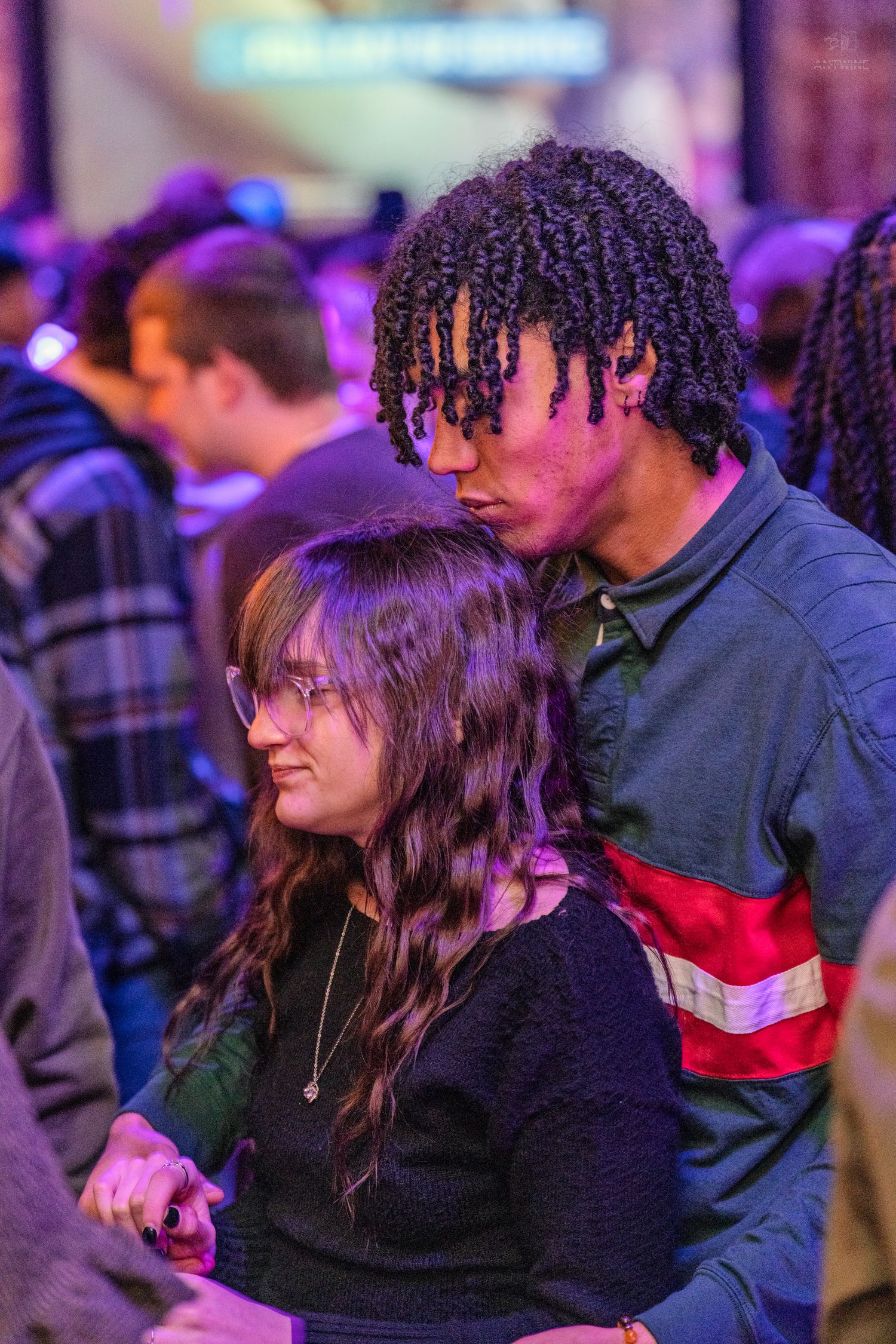 Man with dreadlocks and woman with glasses share a close moment at an indoor event.