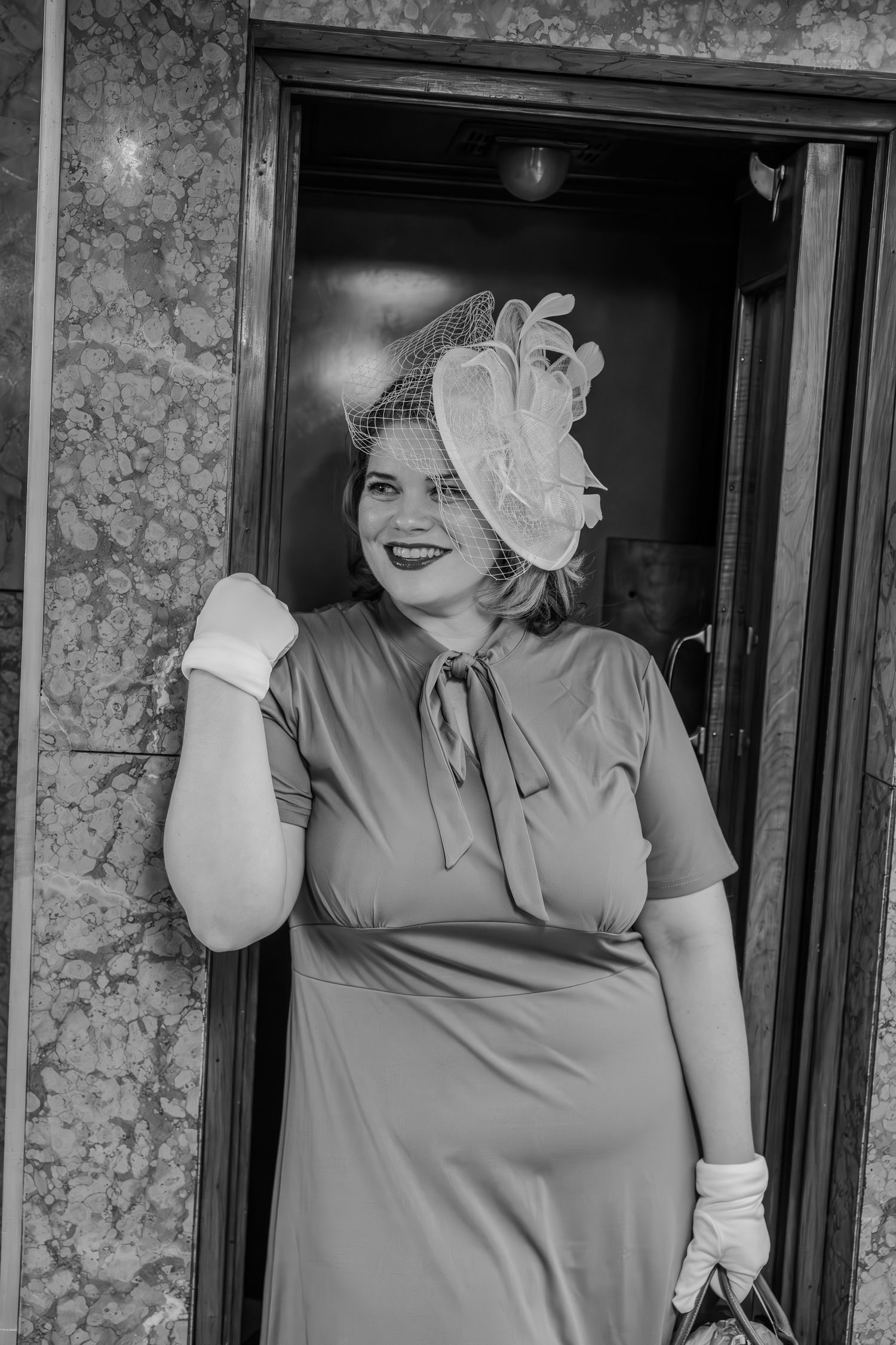 Smiling woman in vintage 1940s dress, fascinator hat, and white gloves posing in doorway, black and white photo.