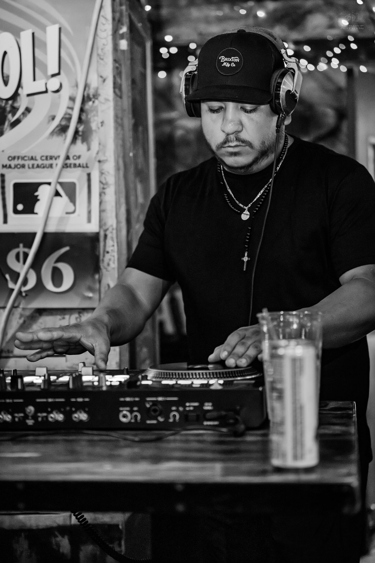 Black and white photo of a DJ wearing headphones and a cap, mixing tracks on turntables at an outdoor venue.