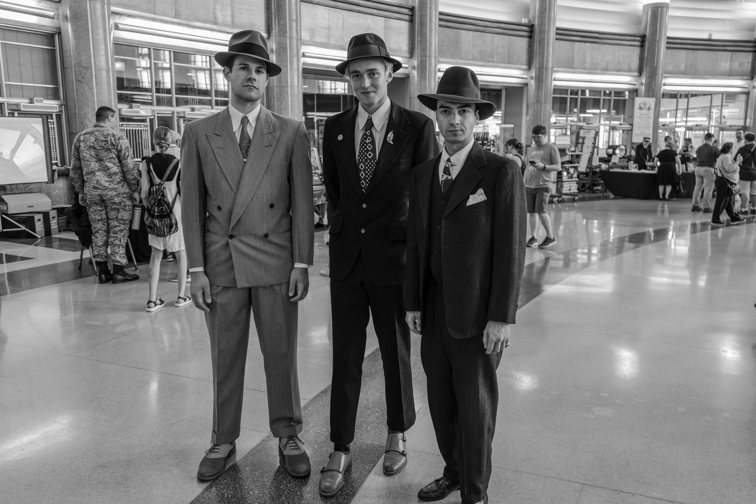 Three men in vintage 1940s suits and fedora hats pose together in a large indoor hall in black and white.