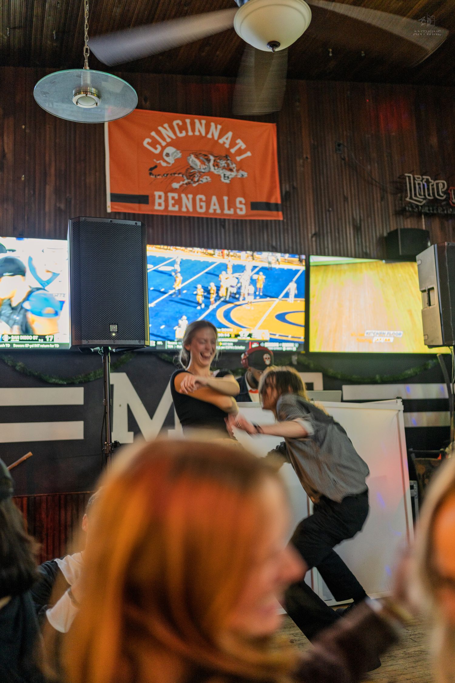 People dancing and socializing inside a bar with a Cincinnati Bengals flag and sports screens on the wall.