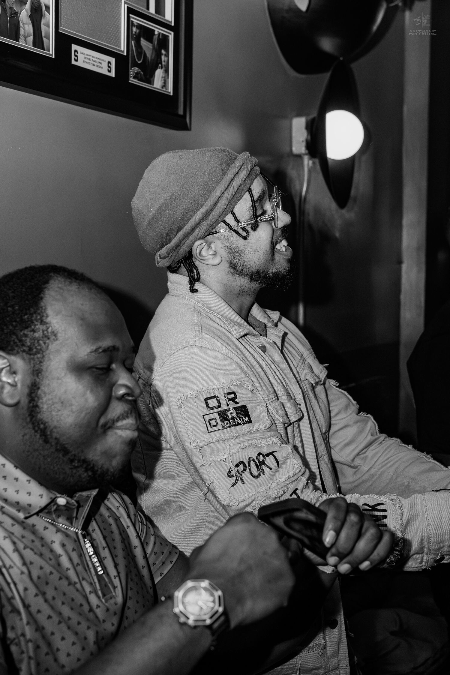 Two men in a dimly lit room, one wearing a beanie and sport jacket, the other a patterned shirt and watch.