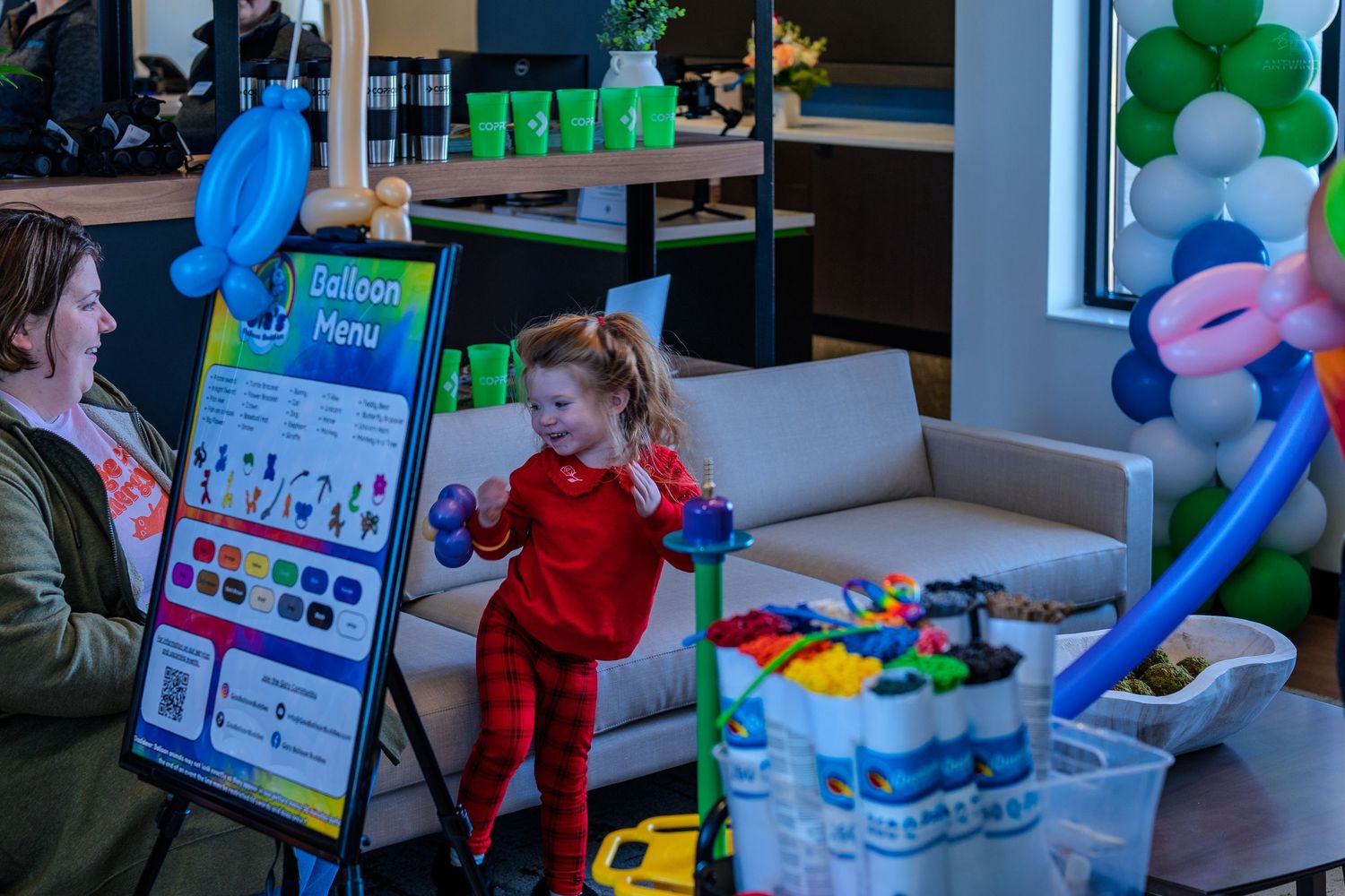 A toddler in red outfit waves near a balloon menu sign at a colorful kids party event indoors.