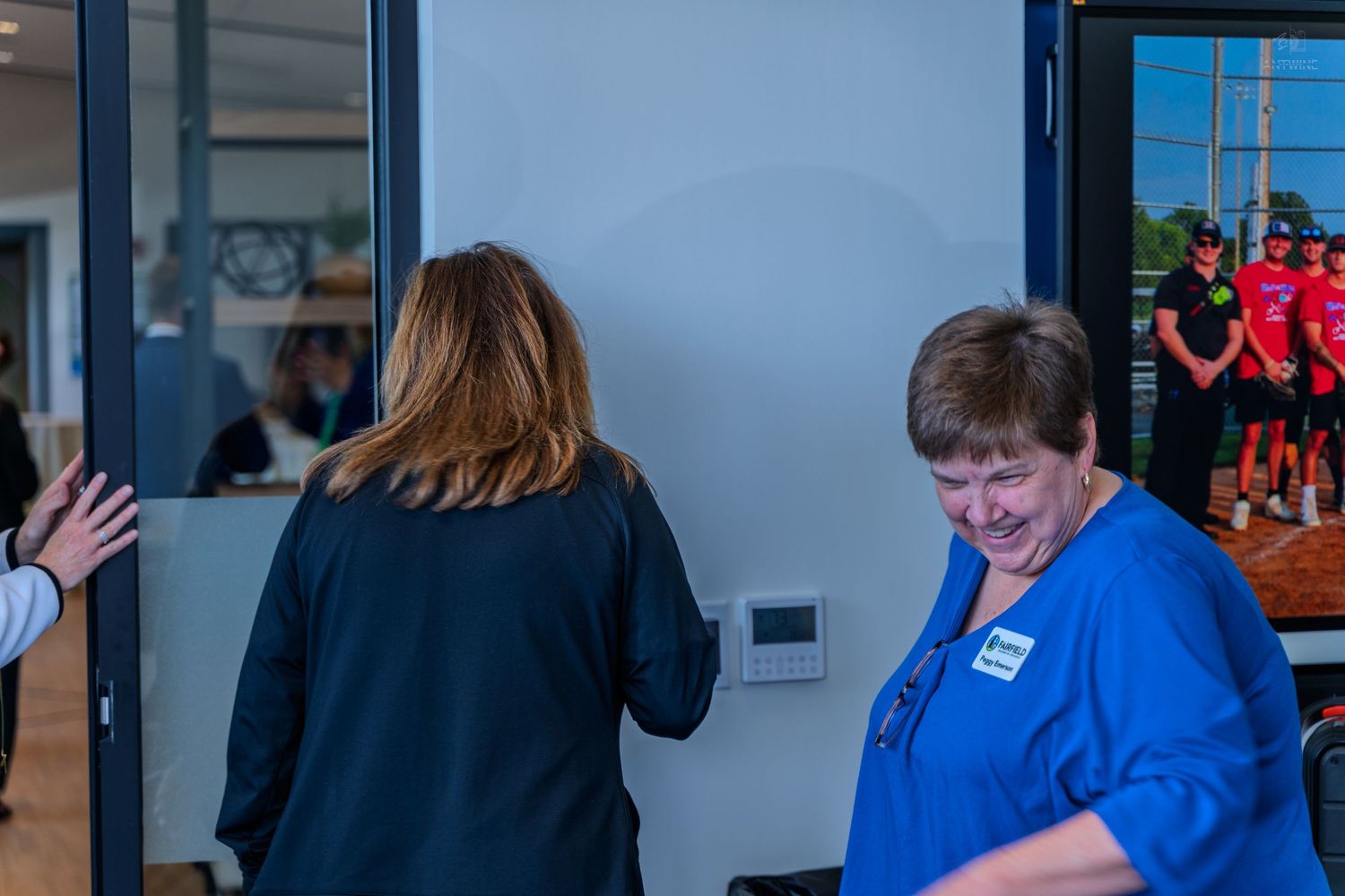 Two women near a glass door, one in blue with a name tag, another in black jacket, with athletes visible through the window.