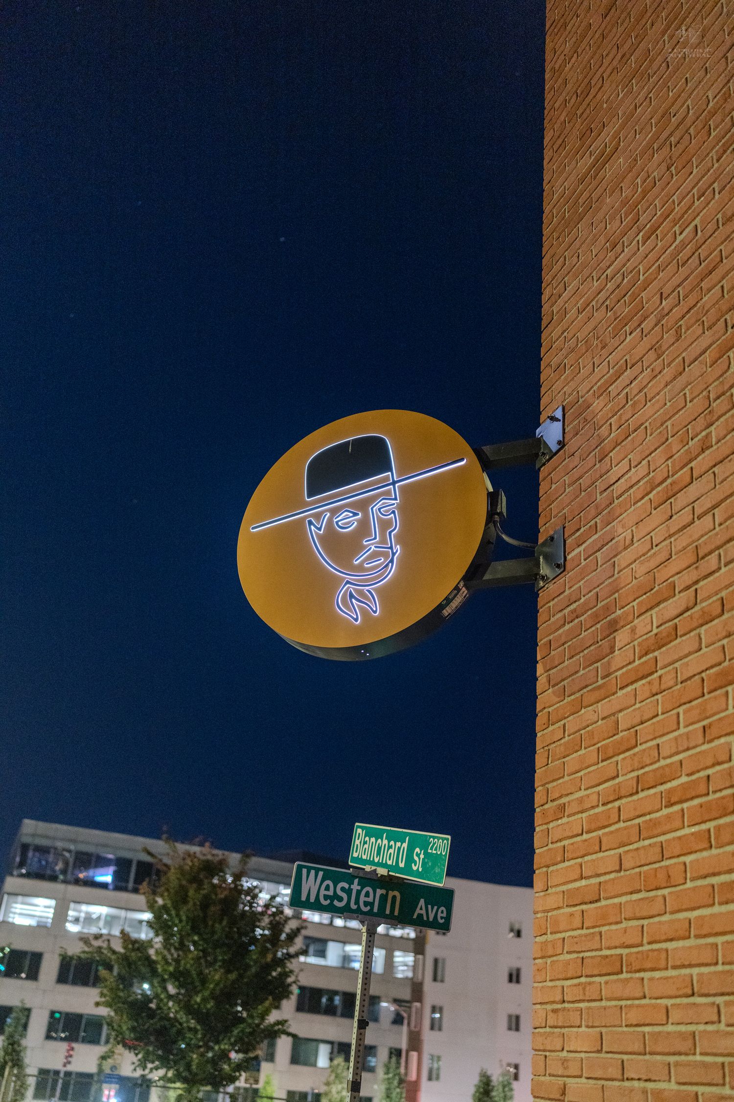 Neon sign of a face with hat on yellow circle at Western Ave and Bamford St corner at night.