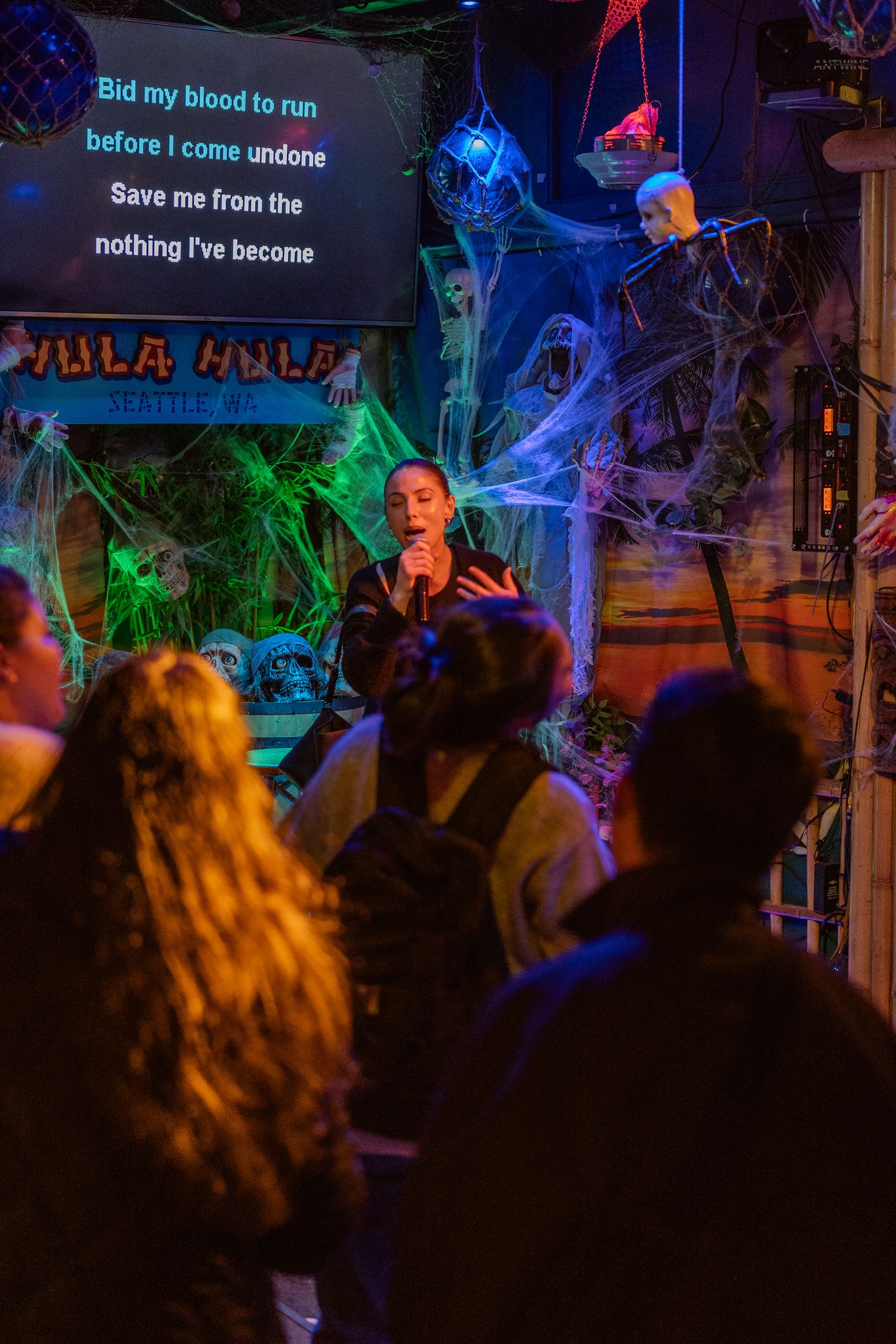 Woman singing karaoke at Hula Hula bar in Seattle with Halloween decorations and spooky cobwebs.