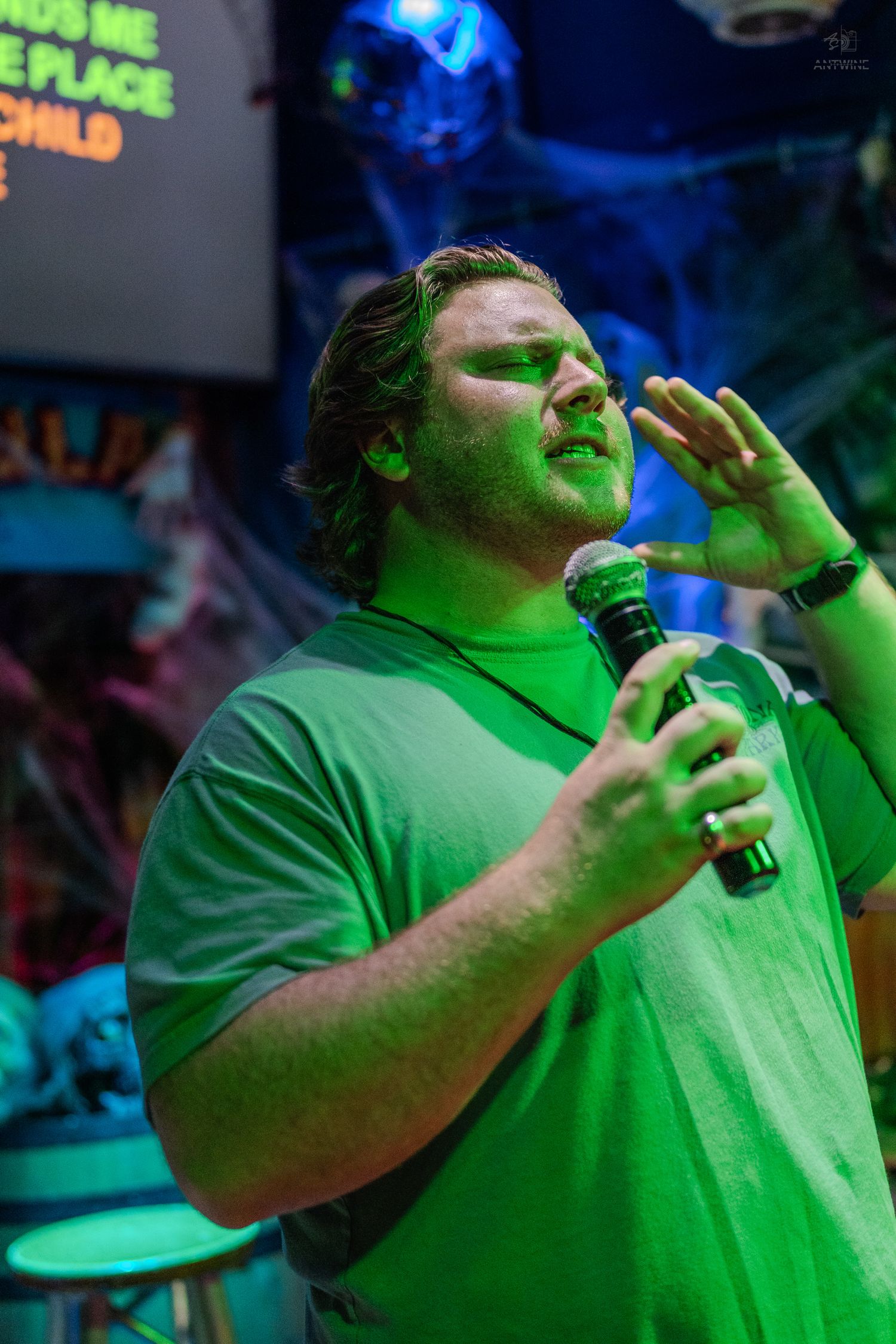 Man in green shirt singing karaoke with microphone under green stage lighting in a decorated bar.