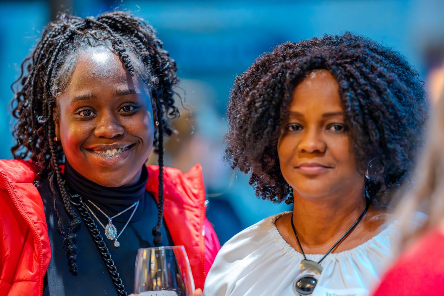 Two women with natural hair smiling at a social event, one in red jacket holding a wine glass.