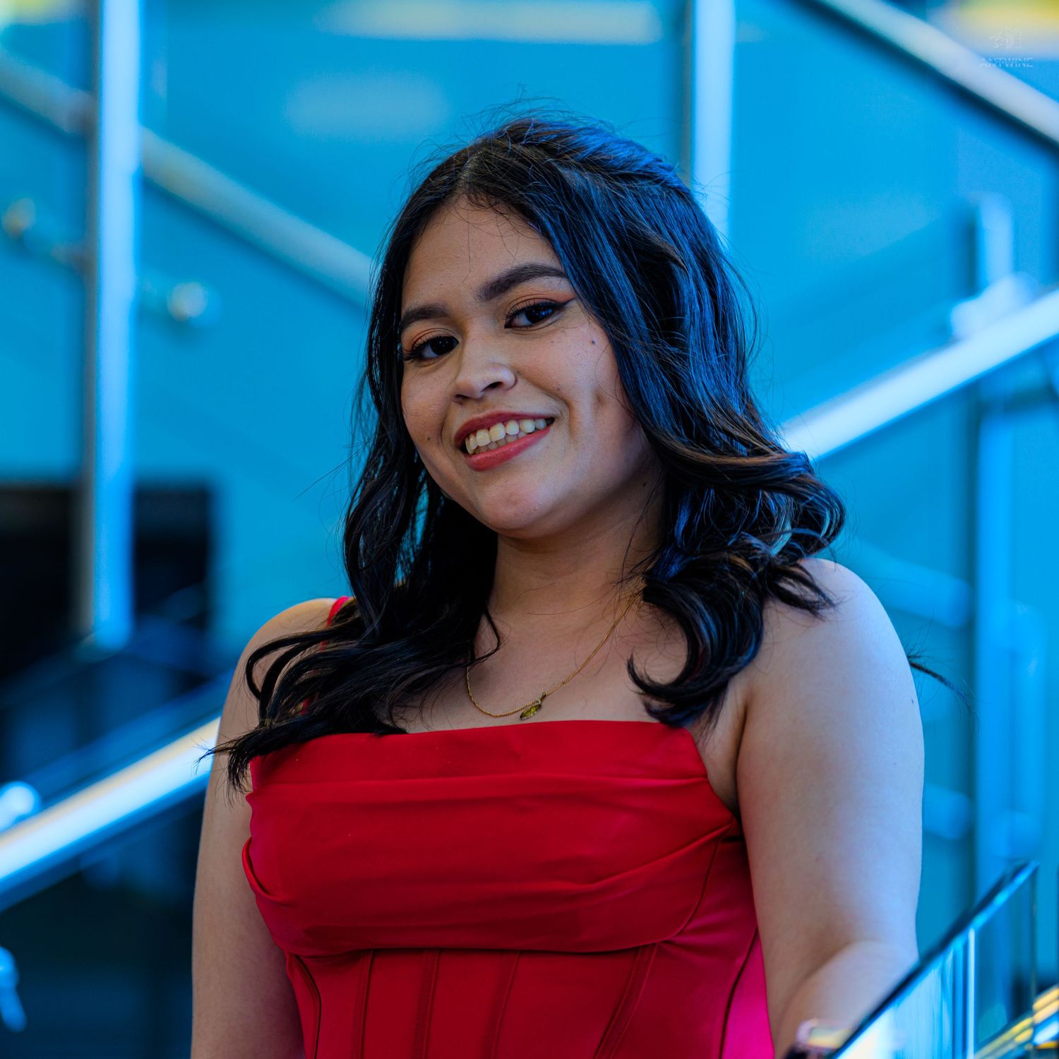 Young woman in red strapless dress smiling on modern staircase with blue glass background.