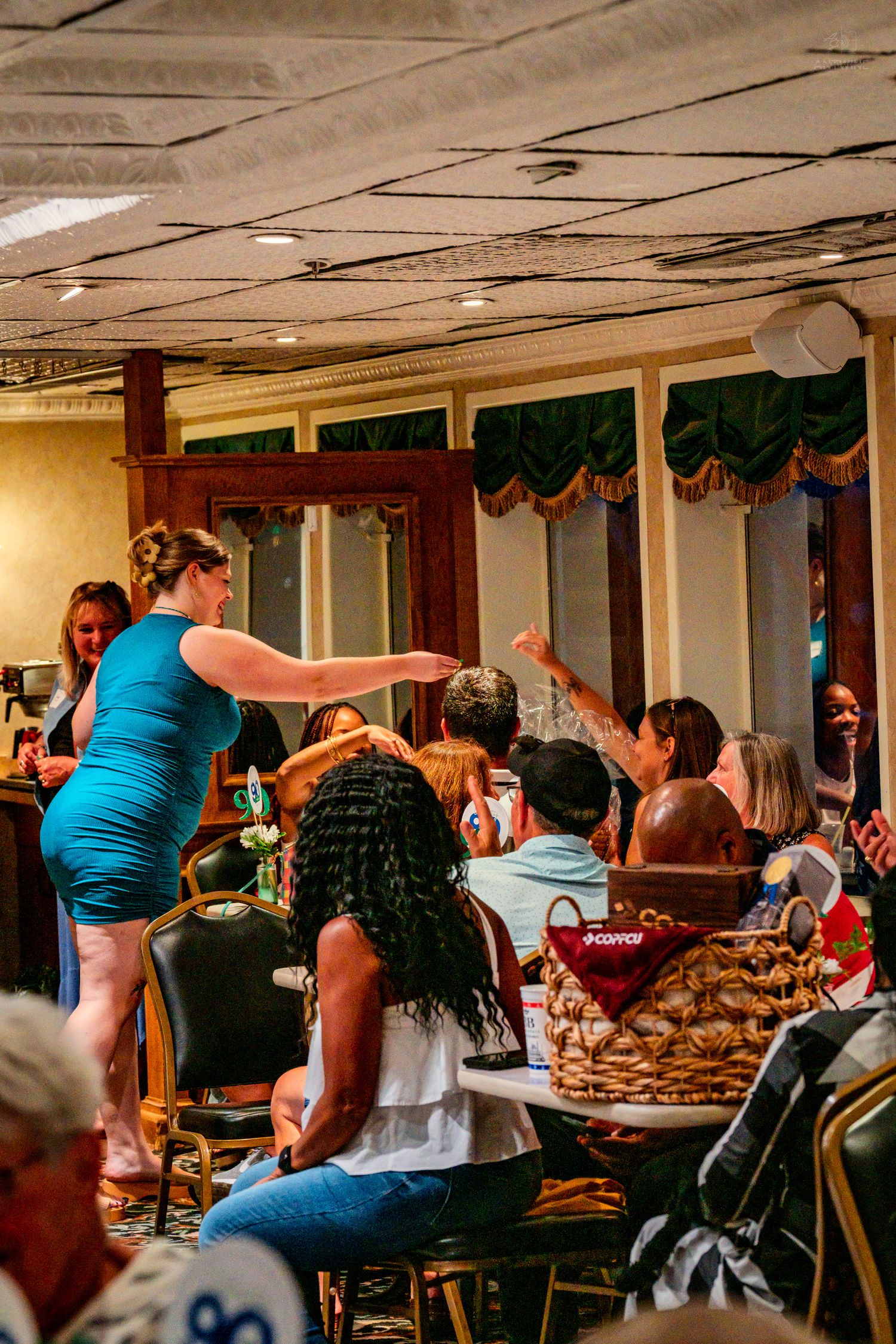 Woman in teal dress points toward excited guests at a lively indoor banquet event.
