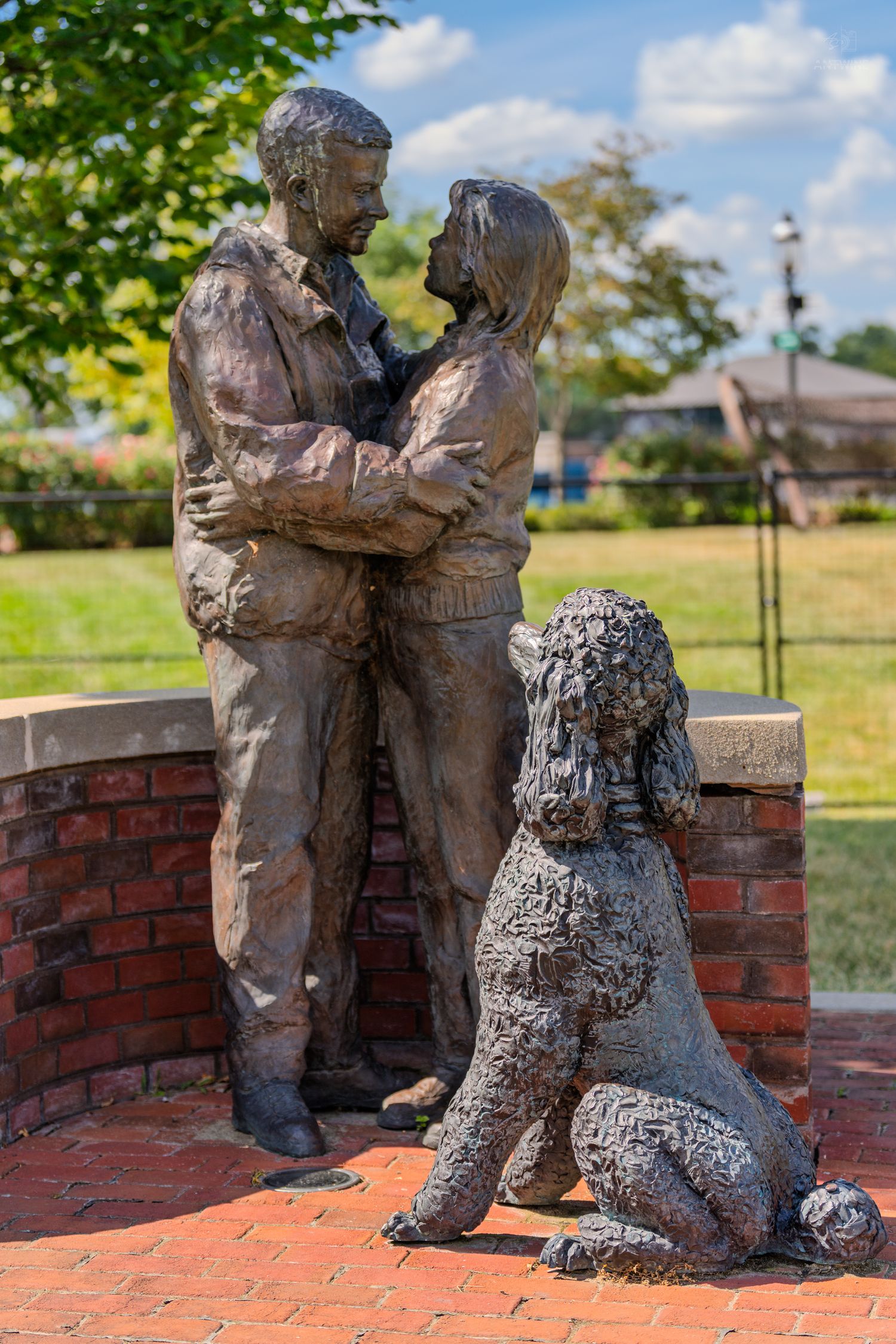 Bronze sculpture of a couple embracing with a poodle sitting beside them on a brick platform outdoors.