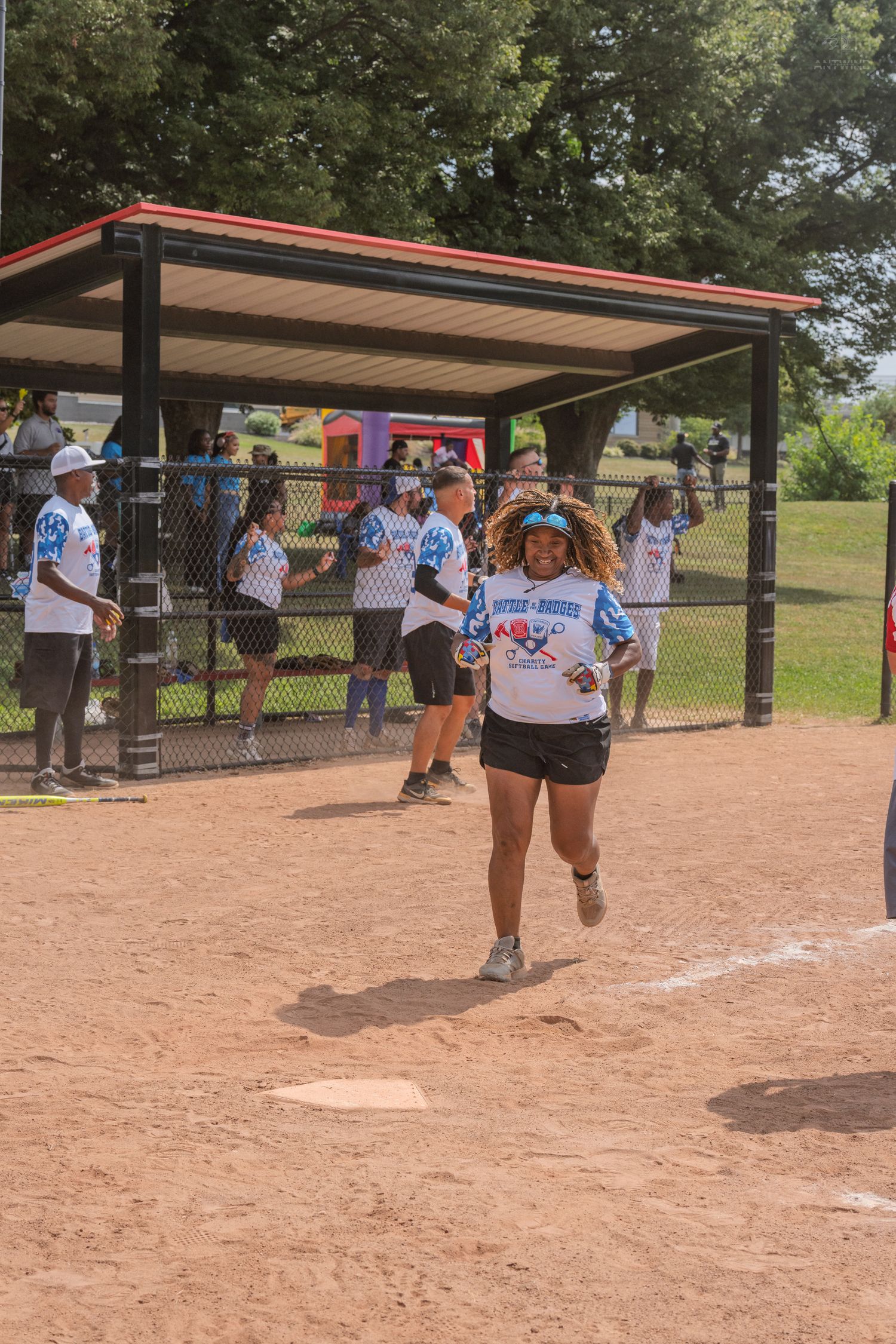 Female softball player runs bases past dugout filled with teammates during charity softball game.