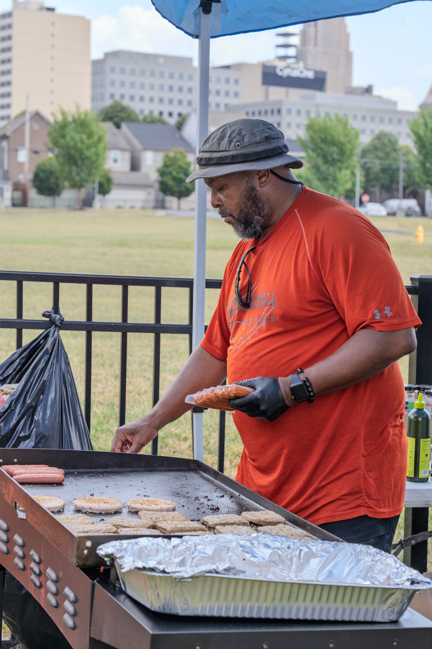Man in orange shirt grilling hot dogs and burgers at an outdoor charity softball game event.