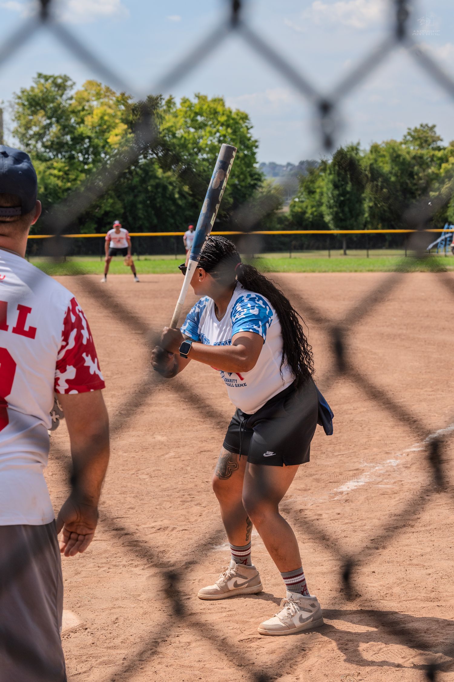 Female softball batter in white uniform ready to swing, photographed through a chain-link fence outdoors.