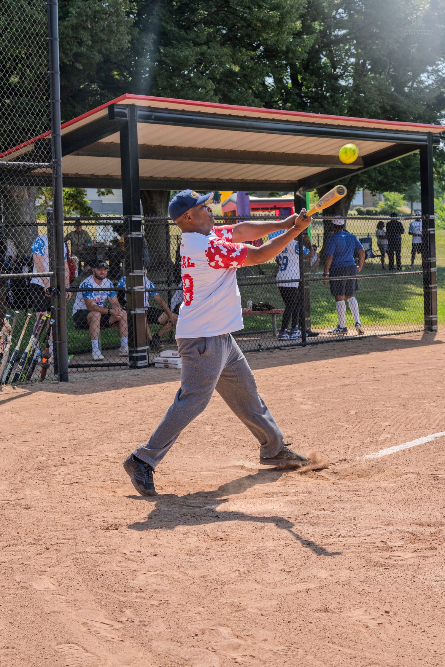 Batter swinging at a yellow softball during an outdoor game on a dirt field with dugout in background.