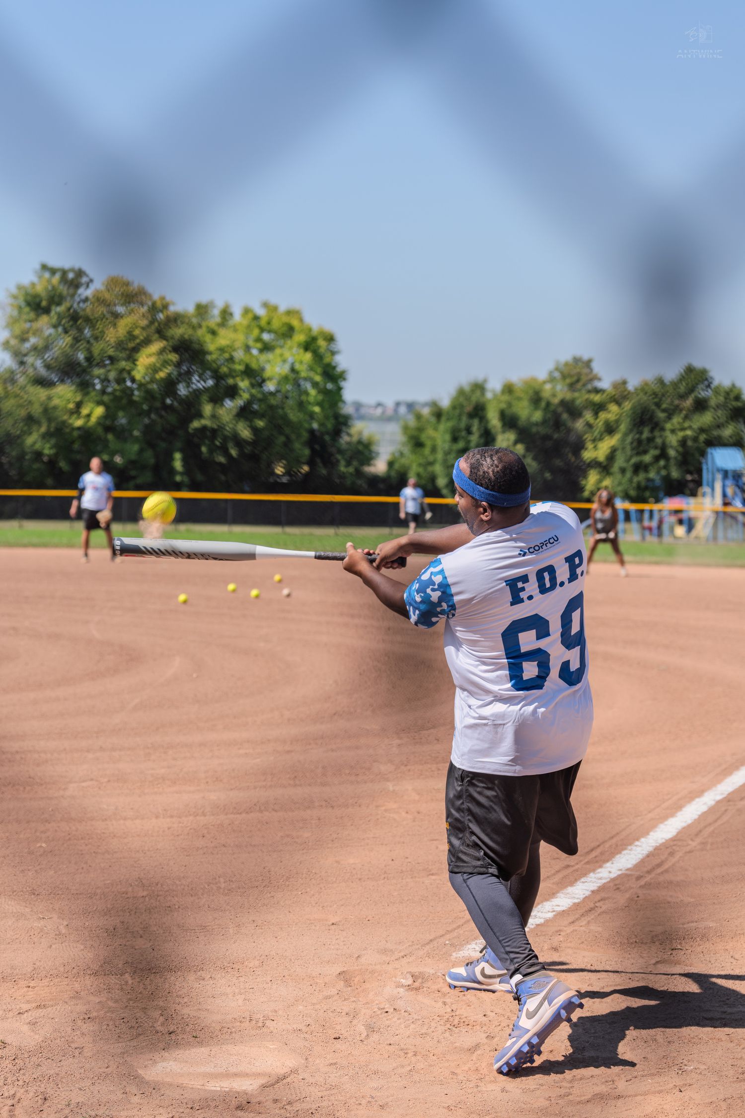 View through fence of softball player in F.O.P. 69 jersey hitting a ball during an outdoor game.