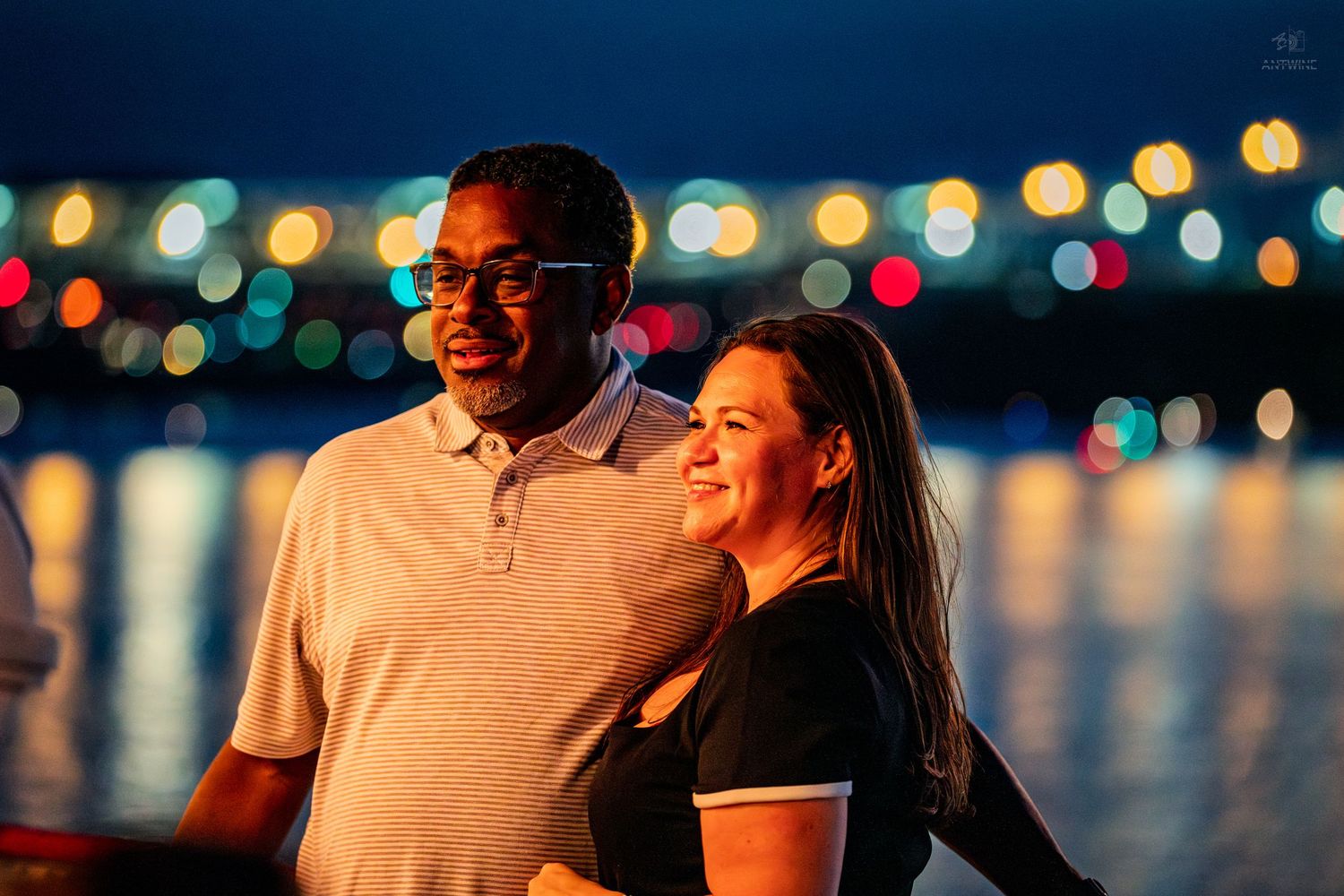 Couple smiling together at night with colorful bokeh city lights reflecting on water behind them.