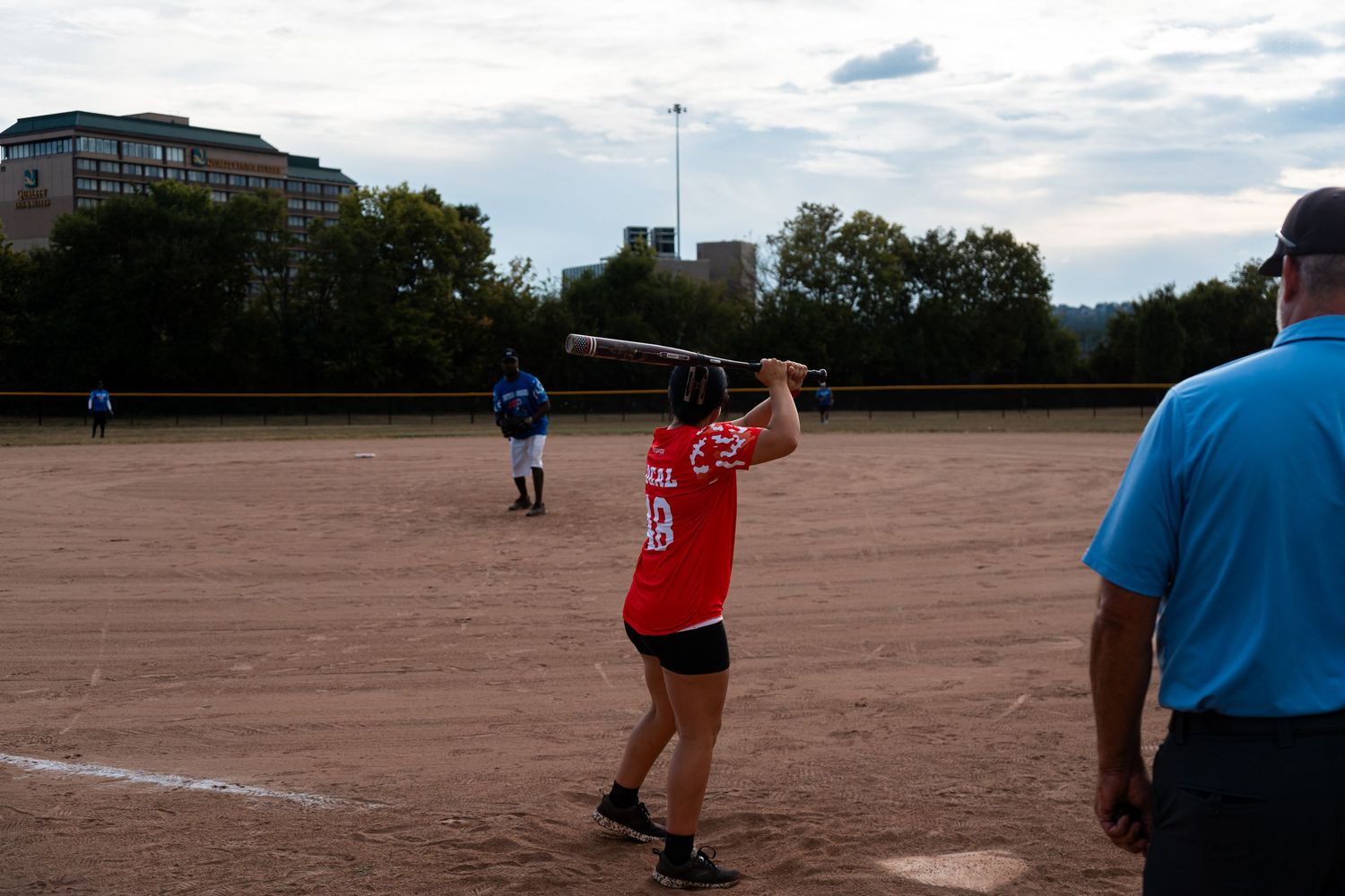 A softball batter in a red jersey swings at a pitch during an evening game with city buildings in background.