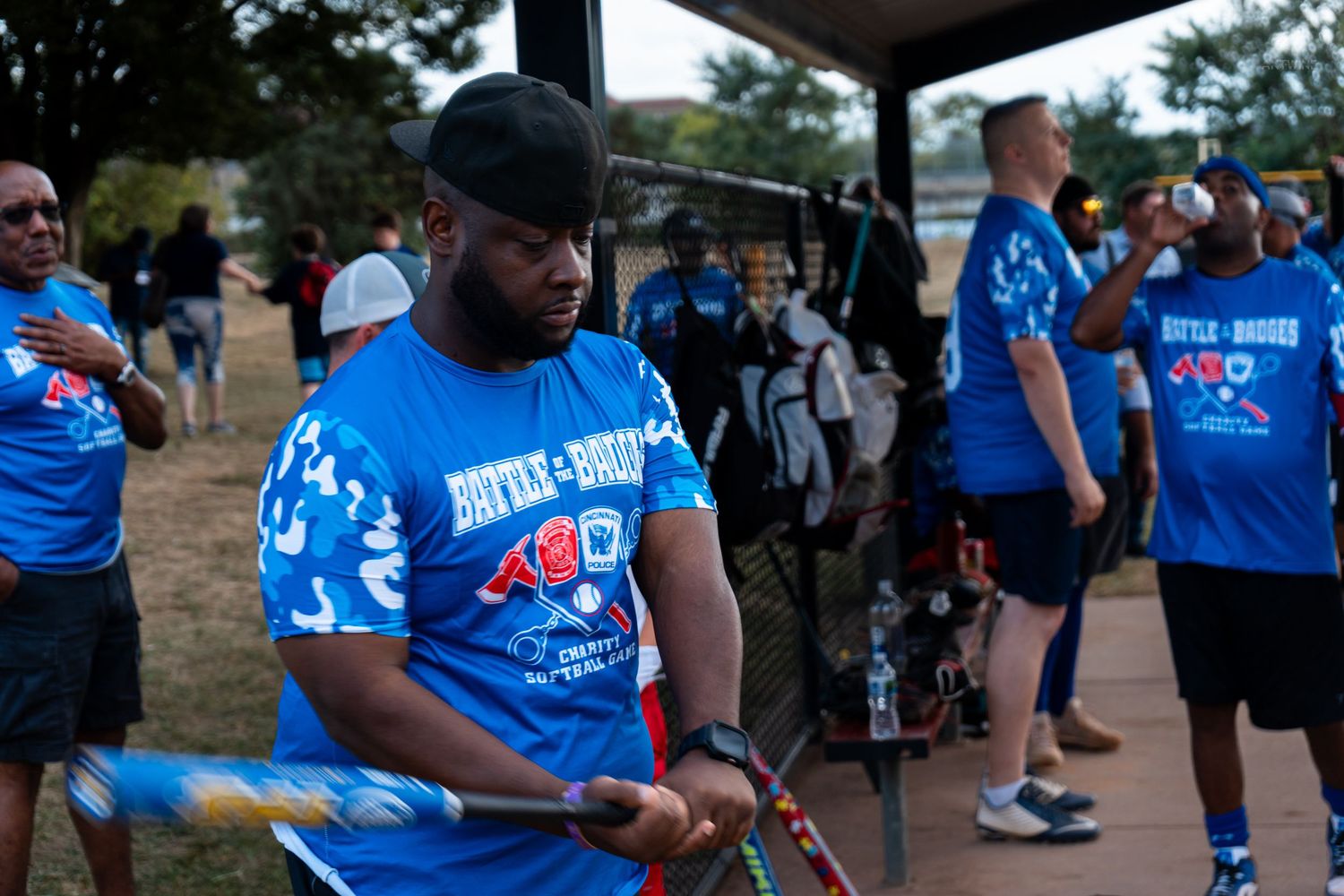 Man in blue Battle-Brothers shirt holds archery equipment at an outdoor charity sports event with participants in matching uniforms.