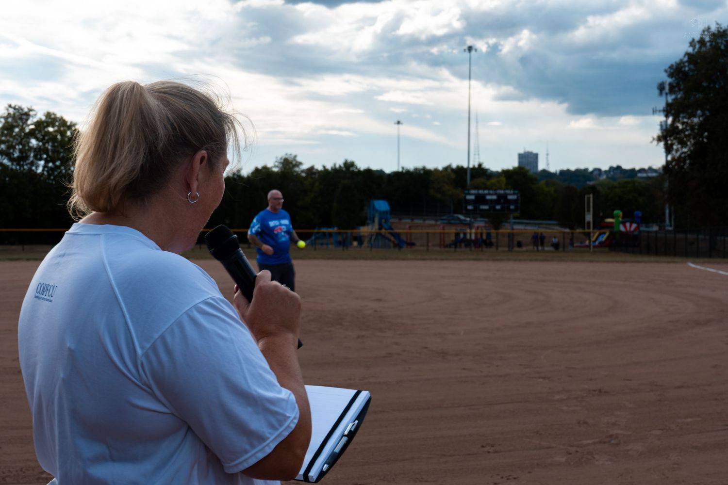 Woman with clipboard observing a softball game on a dirt field at dusk with trees and city skyline in background.