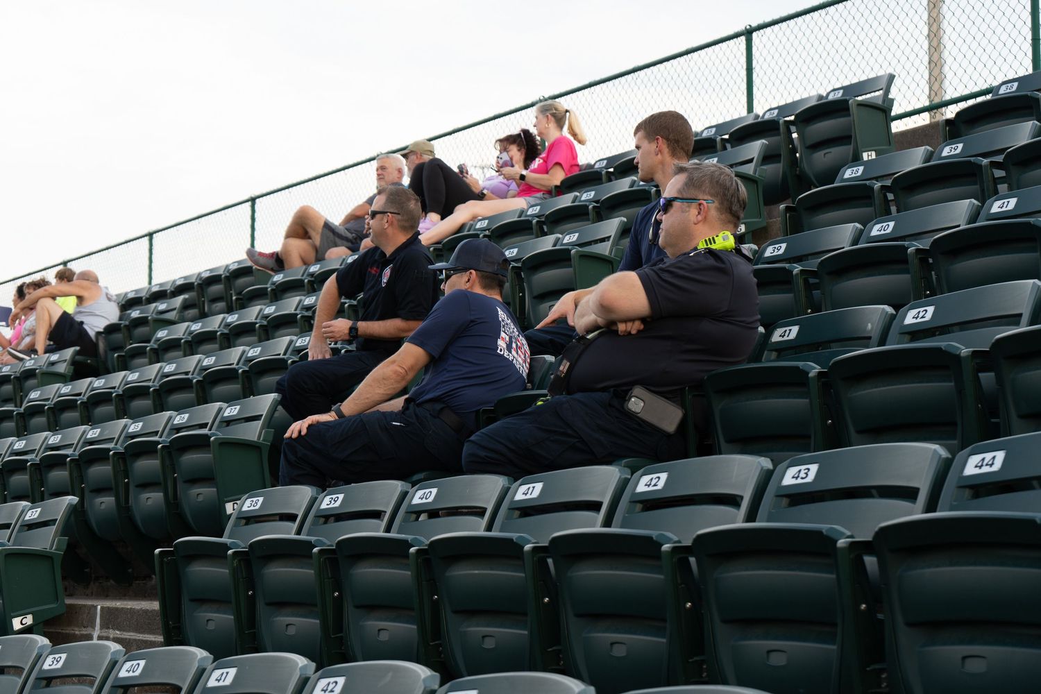 Spectators sitting in dark green stadium bleacher seats watching an outdoor baseball game on a cloudy day.