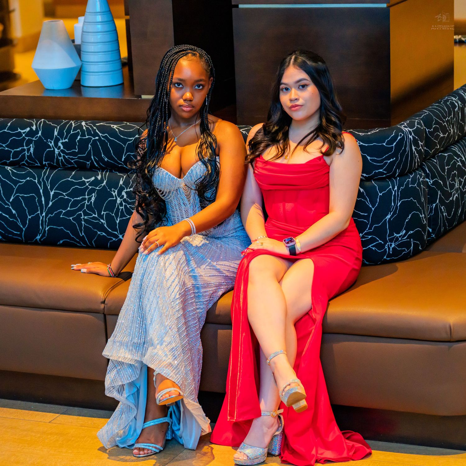 Two young women in formal prom dresses posing on a leather sofa, one in blue sequins and one in red satin.