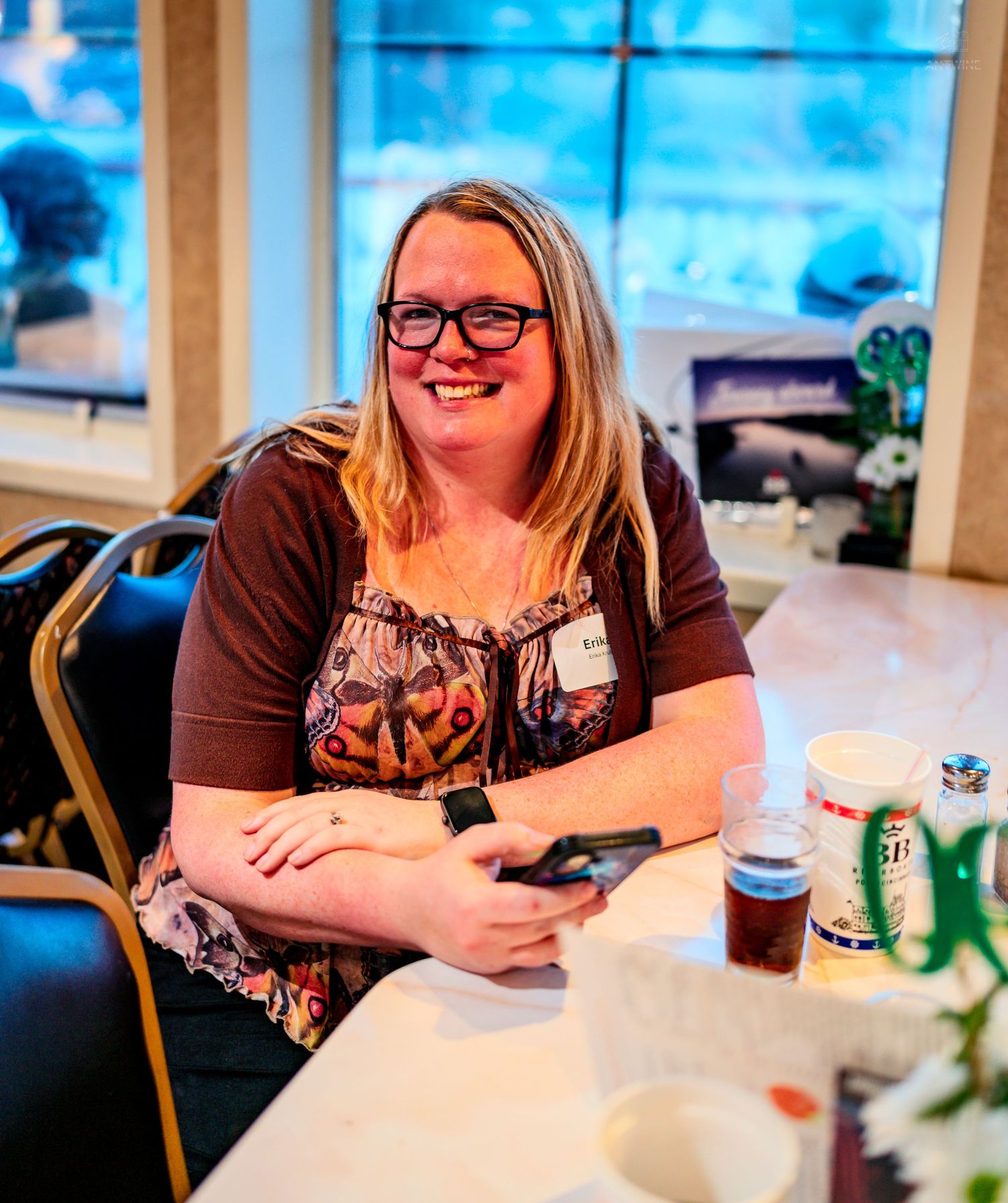 A smiling woman with glasses sits at a banquet table holding a phone during a casual indoor event.